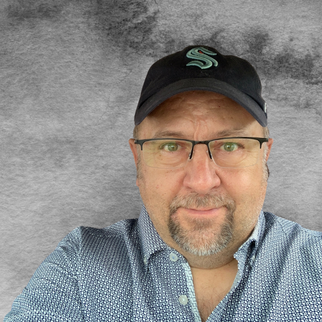 A man wearing glasses, a patterned button-up shirt, and a black baseball cap with a light teal logo, standing in front of a textured gray background.