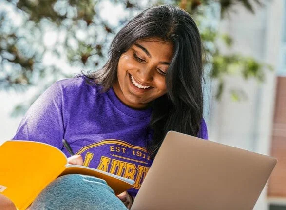 A woman sitting outdoors, smiling and writing in a notebook with a yellow cover, while working on a laptop.