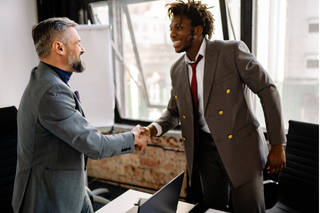 Two men shaking hands in an office, smiling, with large windows and exposed brick wall in the background.