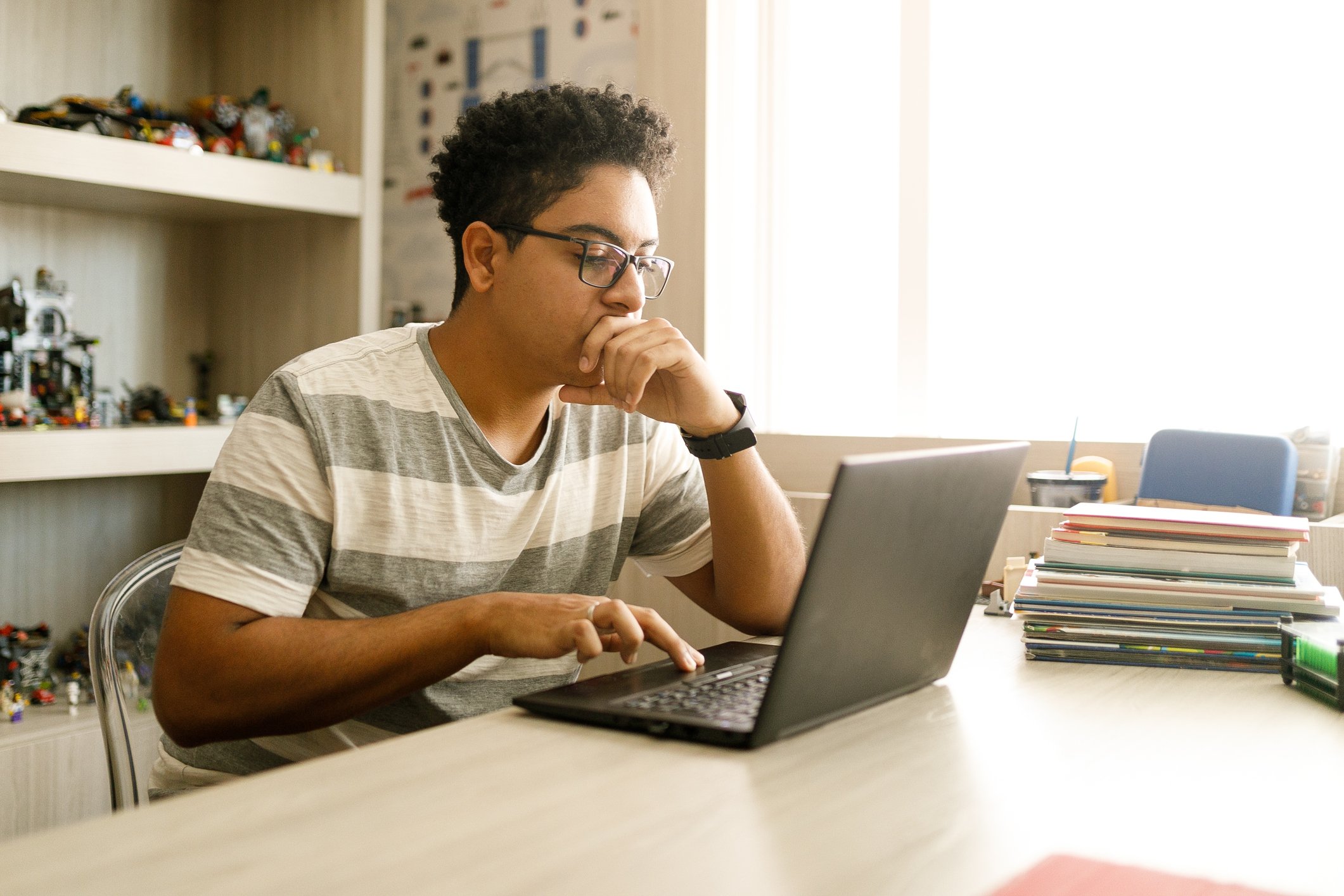 Young man with curly hair and glasses sitting at a desk, looking at a laptop with a thoughtful expression, hand on chin, in a room with books, papers, and shelves of LEGO sets.