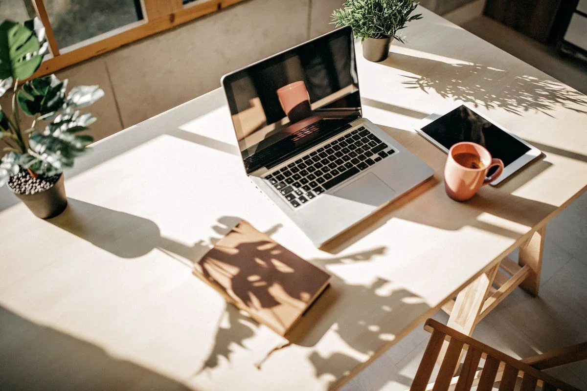A well-lit workspace with a laptop, a tablet, a coffee mug, two potted plants, and a closed notebook on a white wooden table, with shadows and sunlight streaming through a window.