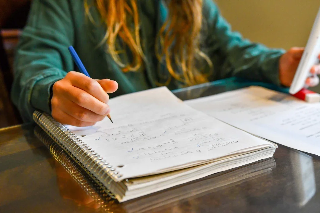 Person writing notes in a notebook on a wooden table, with papers and a smartphone nearby.