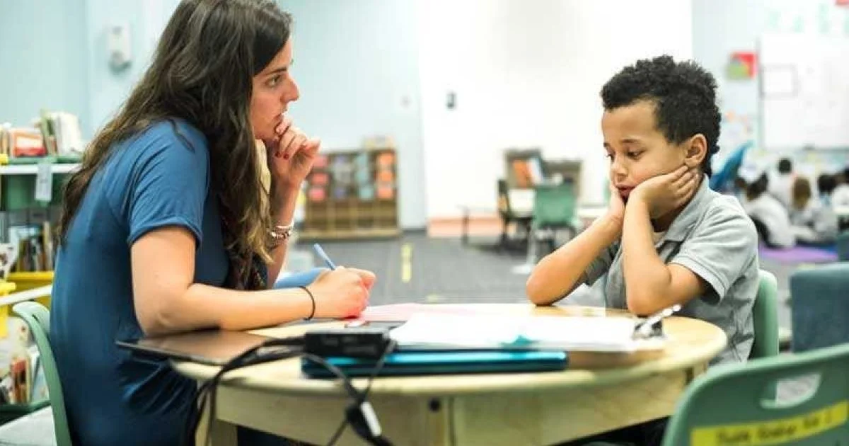 A woman and a young boy sitting at a table in a classroom, with the woman appearing to listen carefully as the boy rests his chin on his hands.
