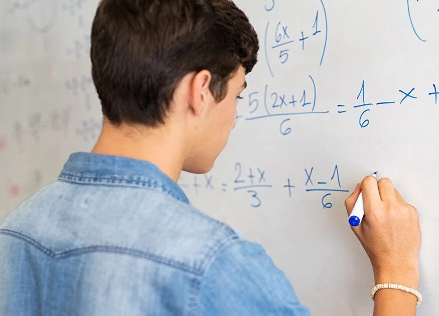 A person wearing a denim jacket solving algebraic equations on a whiteboard using a blue marker.