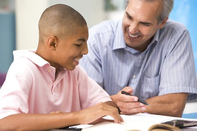 A young boy and an adult man smiling at each other while seated at a table with an open book, indoors.