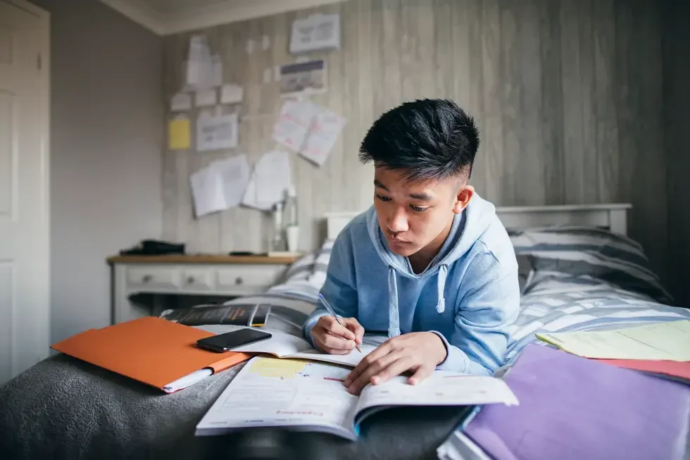 A young boy in a light blue hoodie studies with notebooks, a textbook, and papers spread out on a bed in a bedroom.