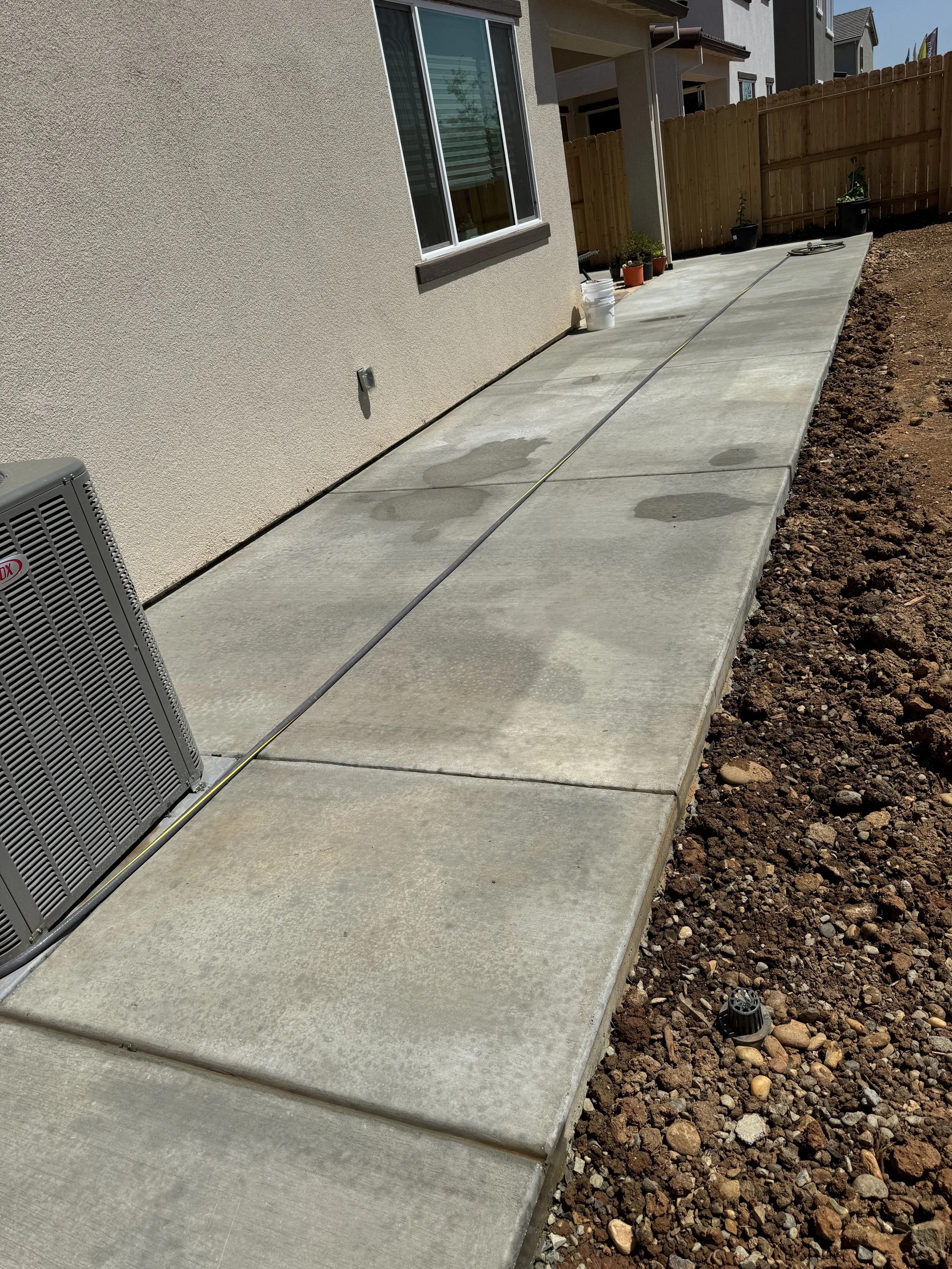 Newly poured concrete patio area beside a house with a window, an air conditioning unit, and nearby potted plants, fenced with wood.