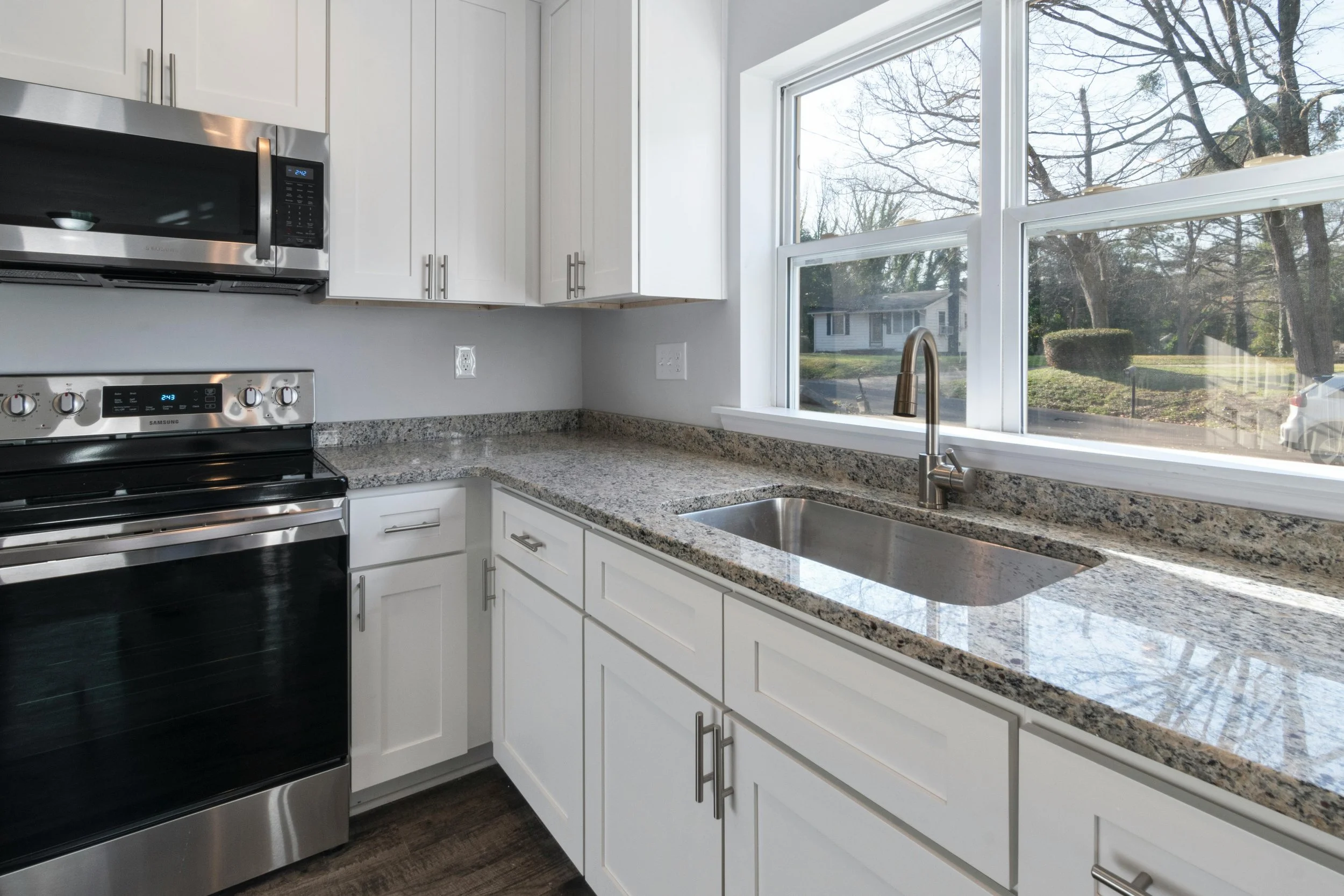 Kitchen with white cabinets, granite countertops, a stainless steel stove and microwave, and a large window overlooking a residential neighborhood.
