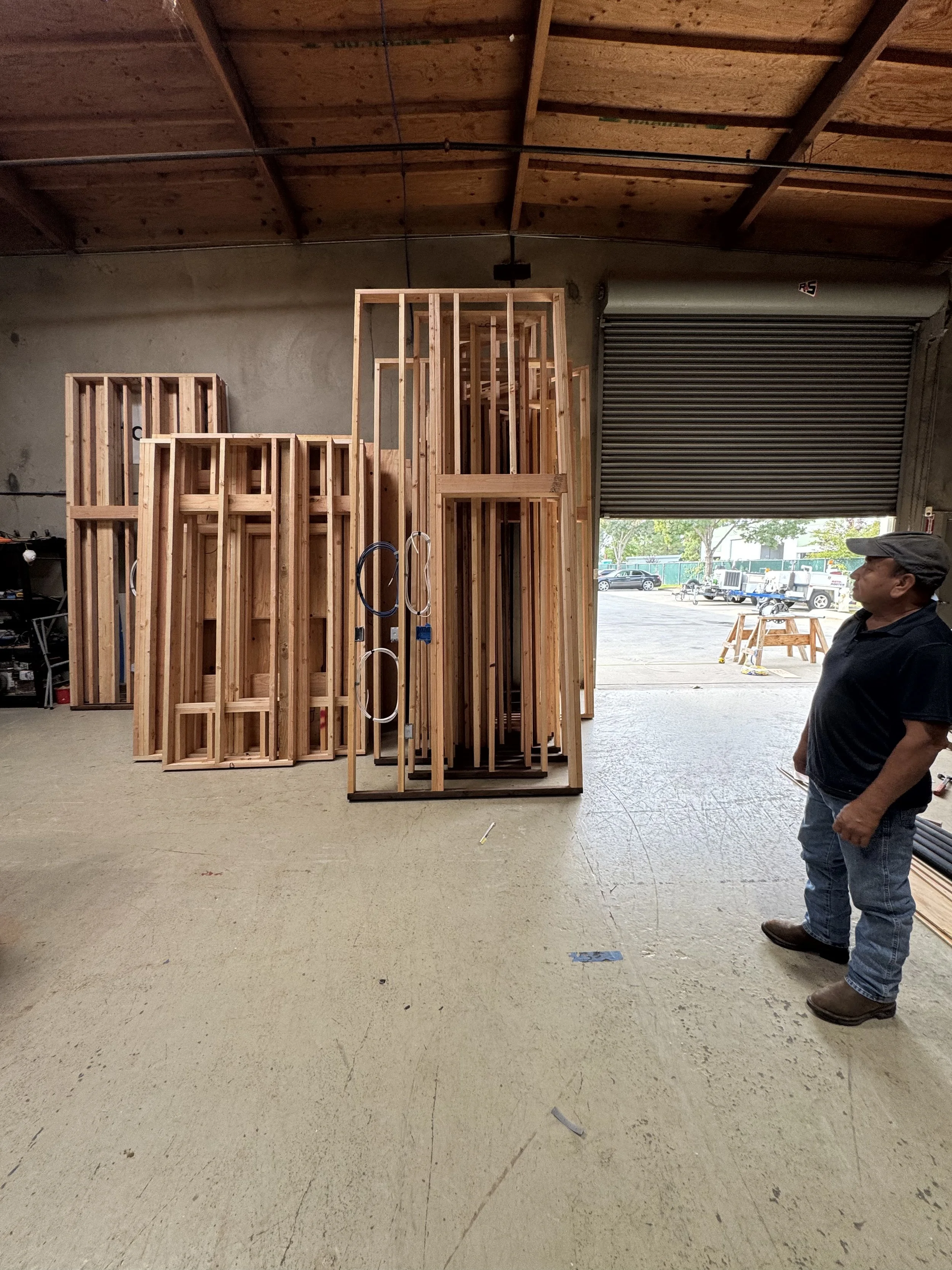 A man standing inside a warehouse observing wooden frames and construction materials, with an open garage door revealing parked vehicles and trees outside.