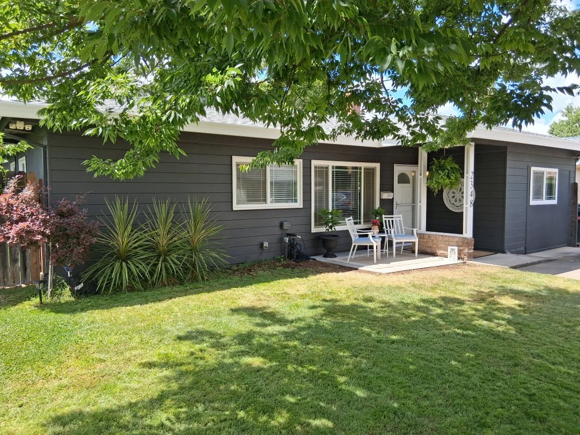 Front yard of a house with green lawn, plants, and trees, showing a black house with white trim, a small porch with patio furniture, potted plants, and a hanging fern, with a large tree overhead providing shade.