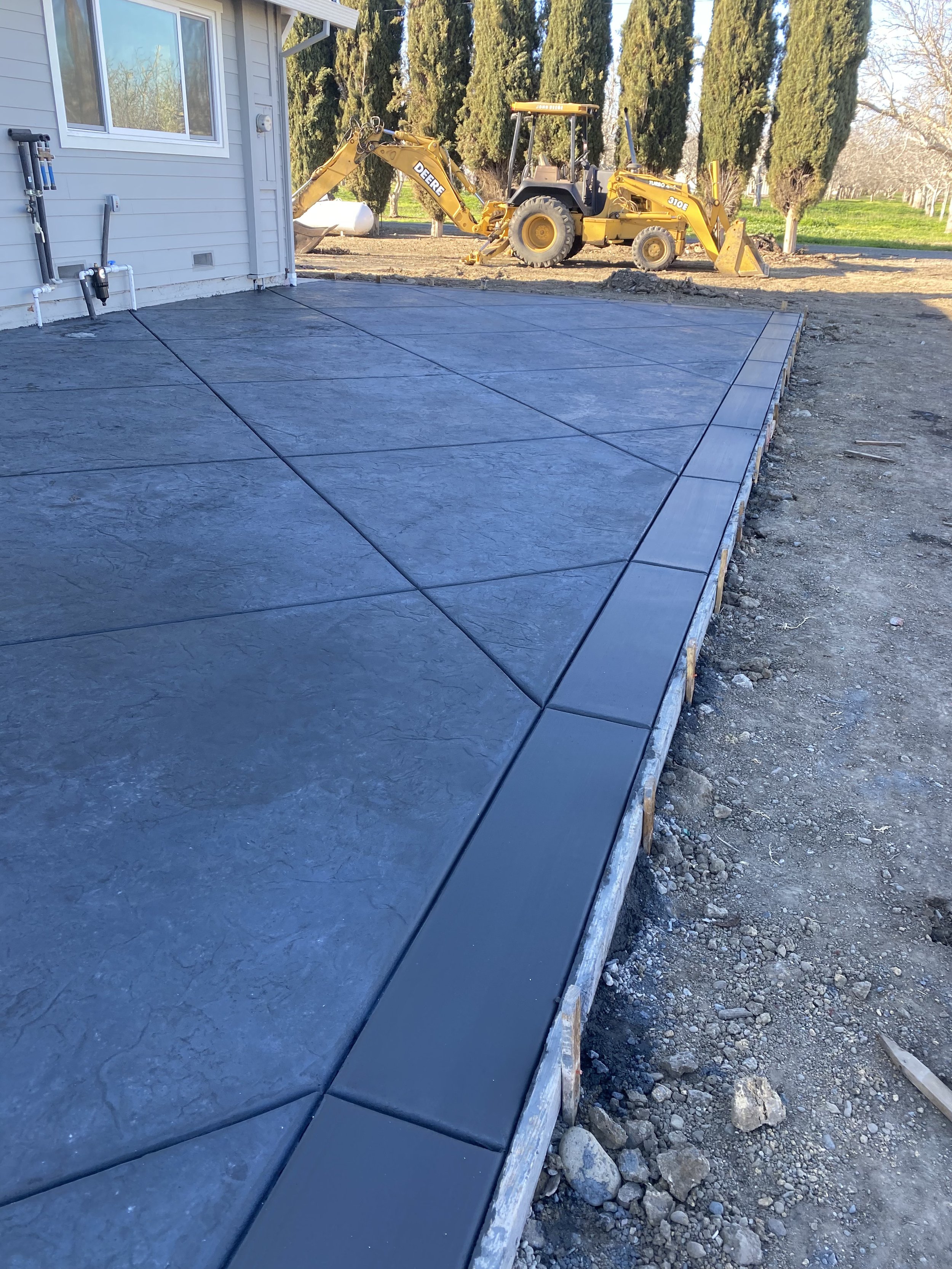 Newly poured concrete patio with decorative border, adjacent to house with a backyard and a yellow backhoe in the background.