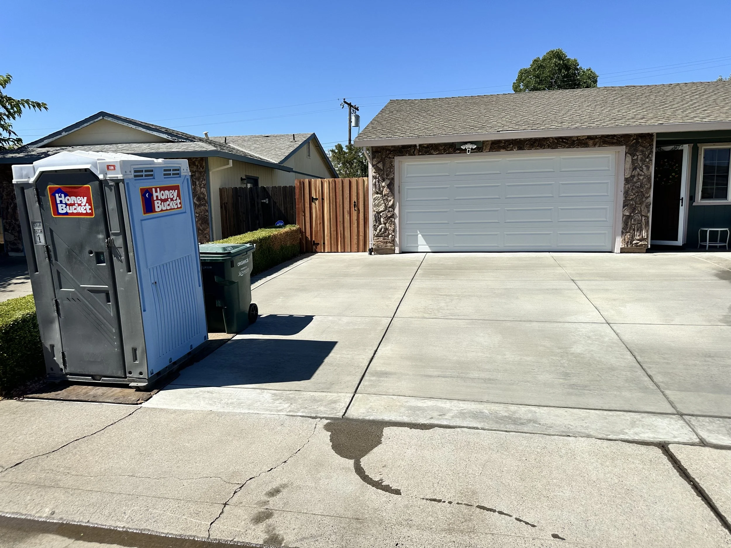 Empty concrete driveway with a large white garage door, a food donation dropbox labeled 'Honey Bucket', and a green recycling bin. A small garden with a hedge and a wooden fence are visible in the background.