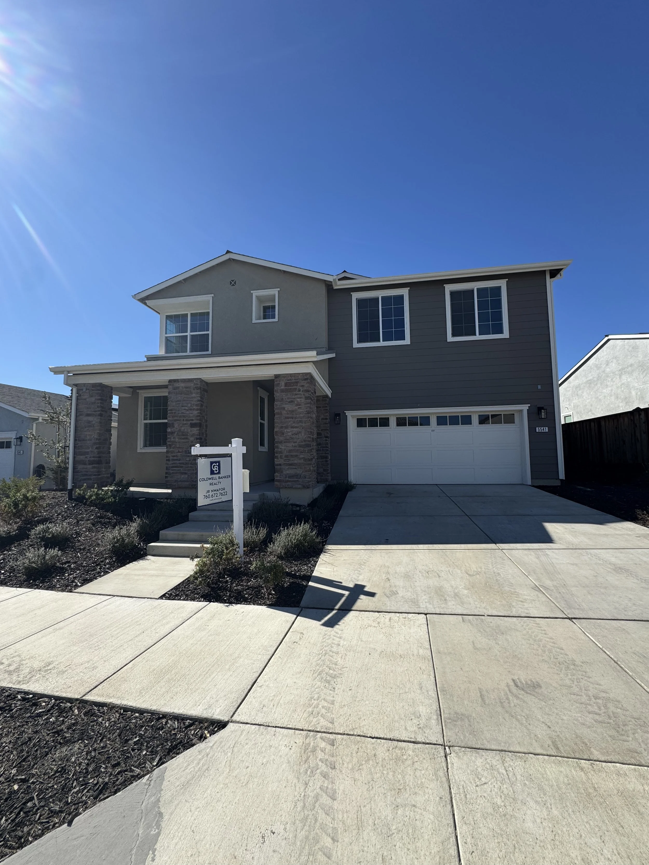 Front view of a modern two-story house with a concrete driveway, stone columns at the porch, and a "For Sale" sign in the front yard. The house has gray and beige exterior walls, multiple windows, and a garage door.