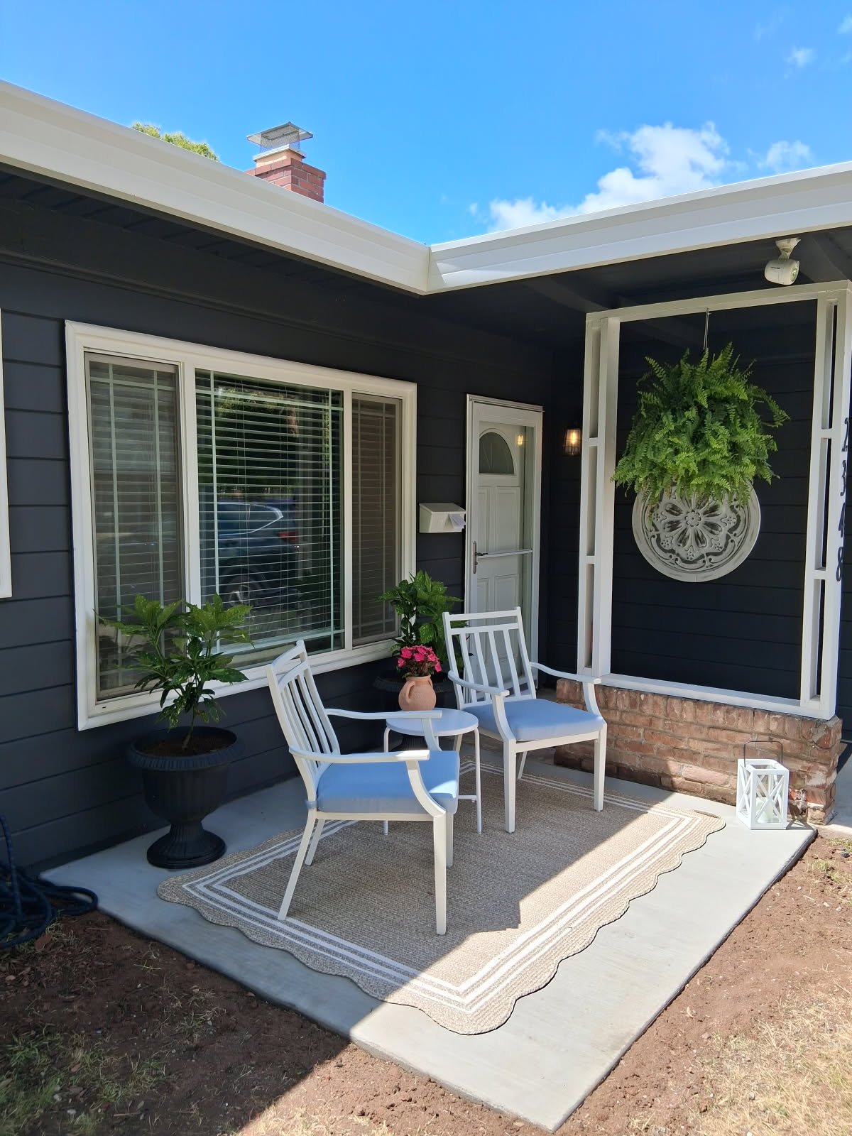 Front porch with two white chairs, potted plants, colorful flowers, a beige rug, and a hanging fern in a decorative wall planter.