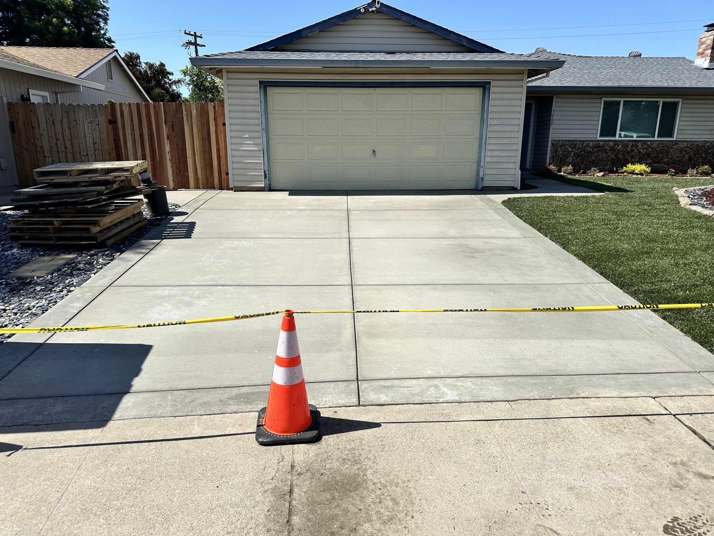 Empty driveway with orange traffic cone and caution tape, leading to a garage on a house with a fenced side yard and a small lawn area.