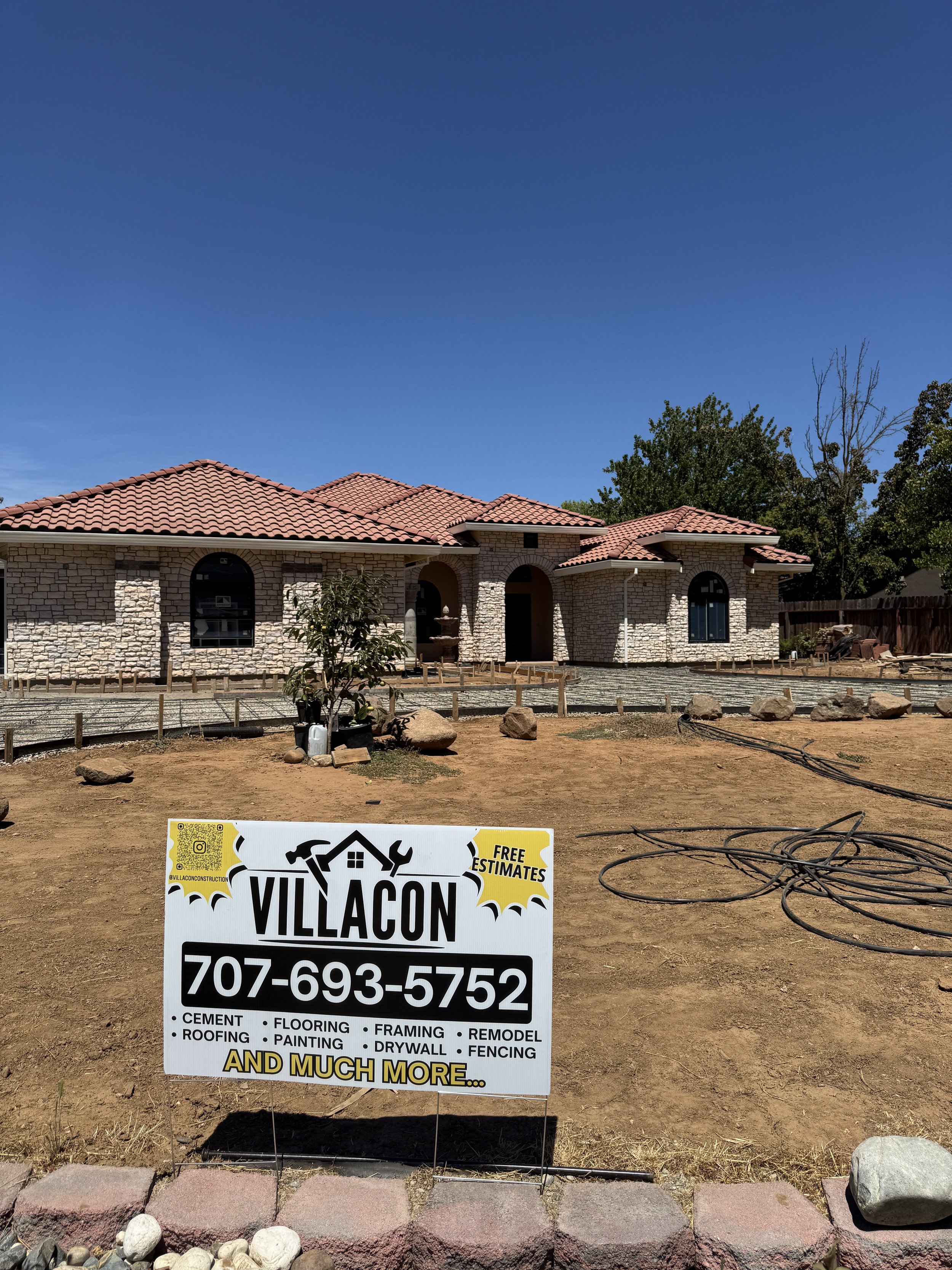 A house under construction with a pink tile roof and stone walls. Construction materials and tools are scattered around the yard. A sign in the foreground displays the name 'VILLACON,' a phone number, and services offered such as cement, roofing, flo