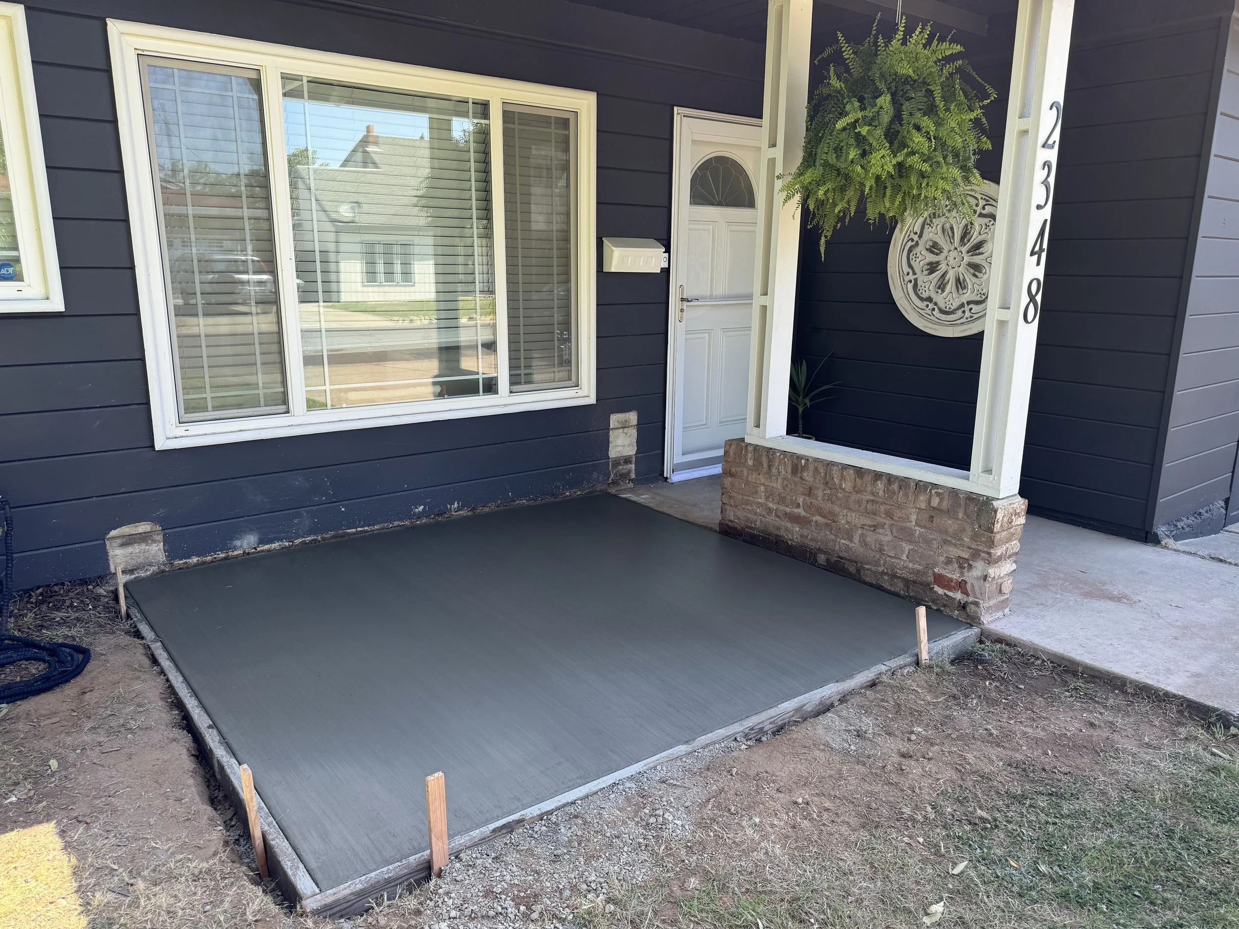 Front porch of a house with a large window, white door, and a newly poured concrete step. The house has dark blue siding, and there is a hanging fern plant and decorative wall art near the entrance.