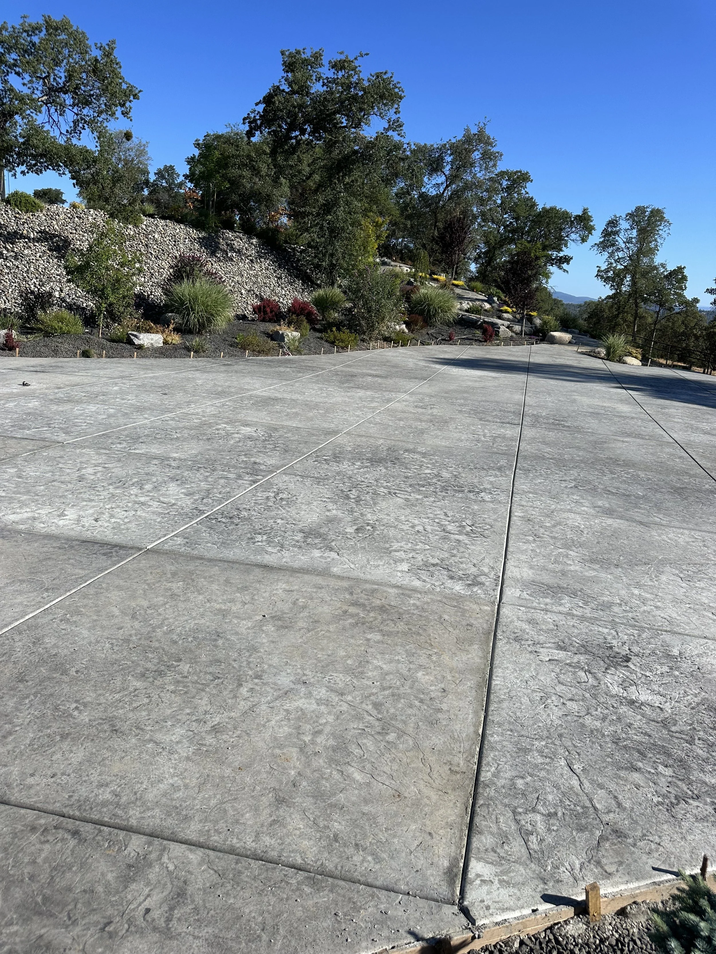 A paved outdoor area with large stone tiles, bordered by landscaped hillside with rocks, bushes, and trees, under a clear blue sky.