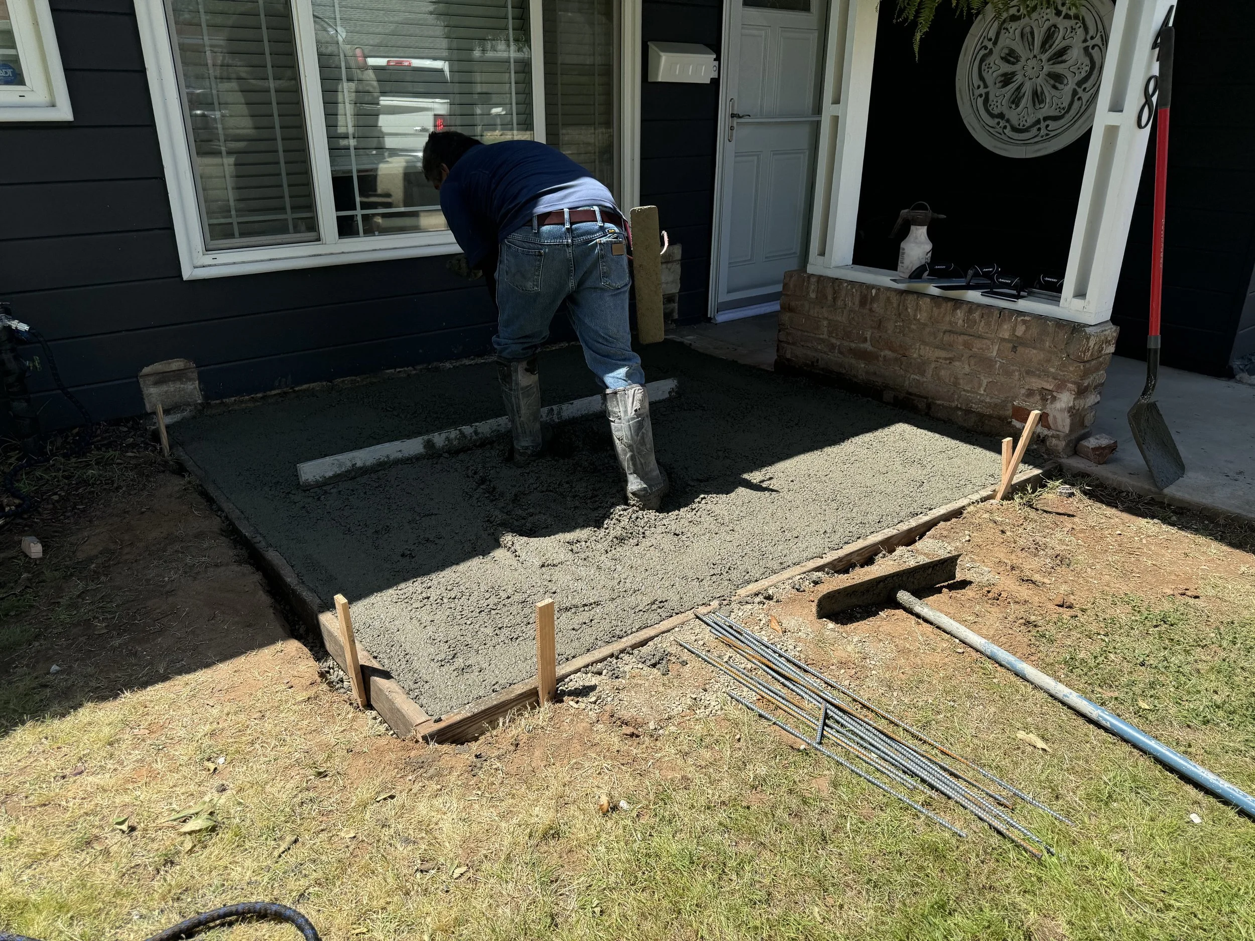 A person is pouring concrete into a small, rectangular outline for a new patio or walkway in front of a house. They are wearing jeans, a dark shirt, and rubber boots. Construction tools and materials are nearby, including a shovel and several steel r