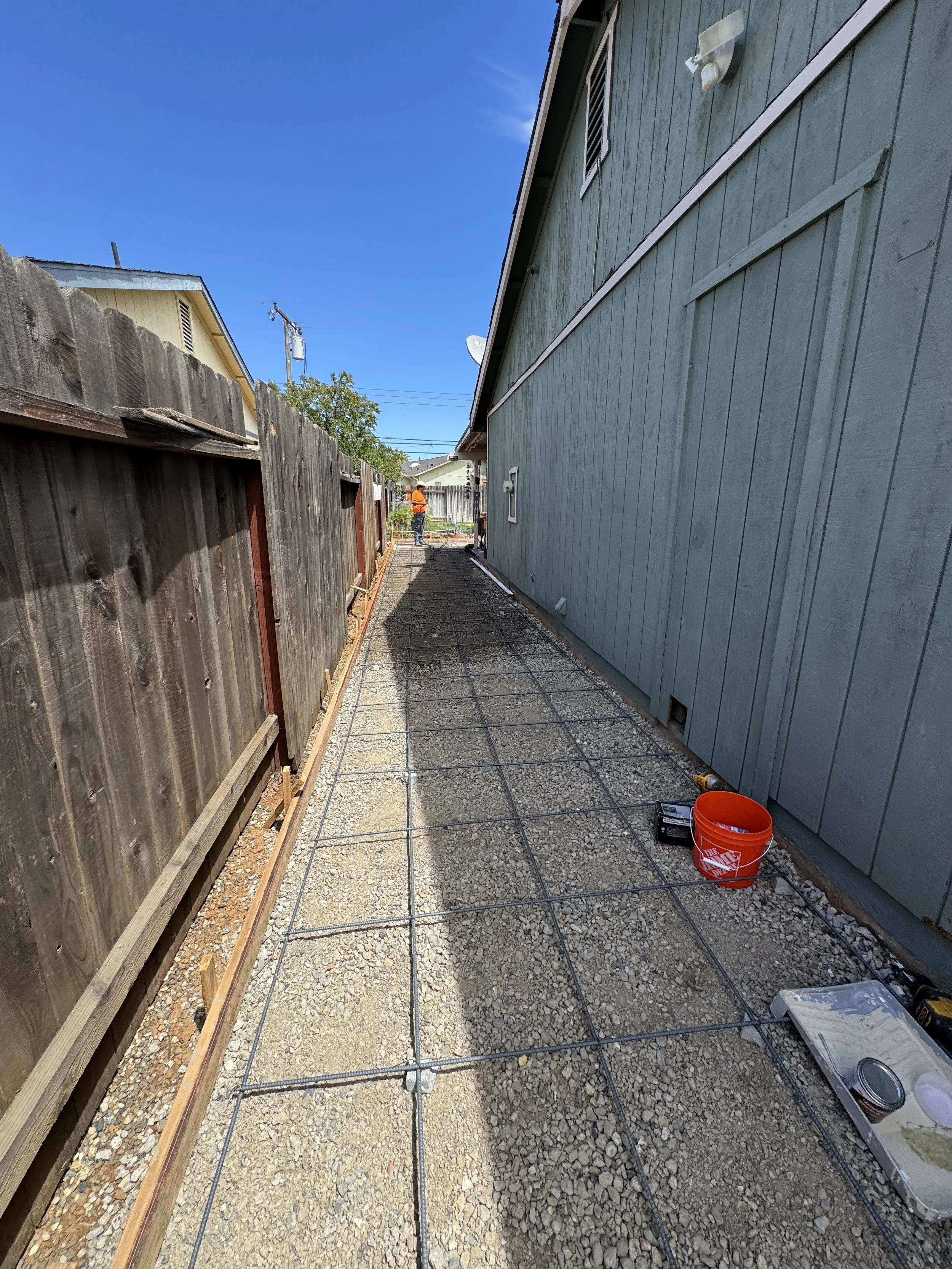 A backyard construction site with a gravel pathway being prepared with a metal grid for a concrete pour. On the right is a blue house with a window and utility boxes, and on the left is a weathered wooden fence. A worker in an orange shirt and hard h
