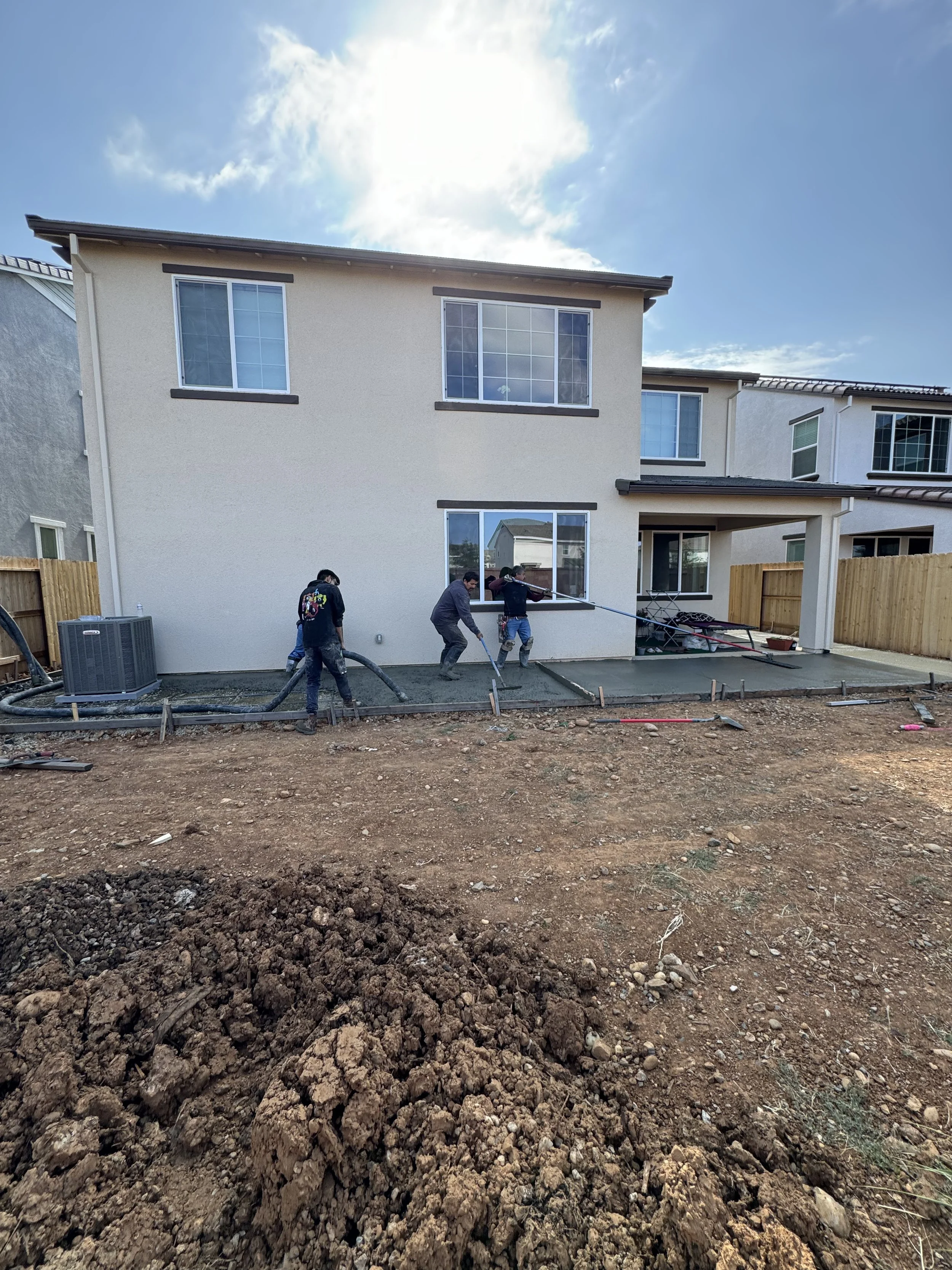 Workers laying concrete on a backyard patio in front of a two-story house under a partly cloudy sky.