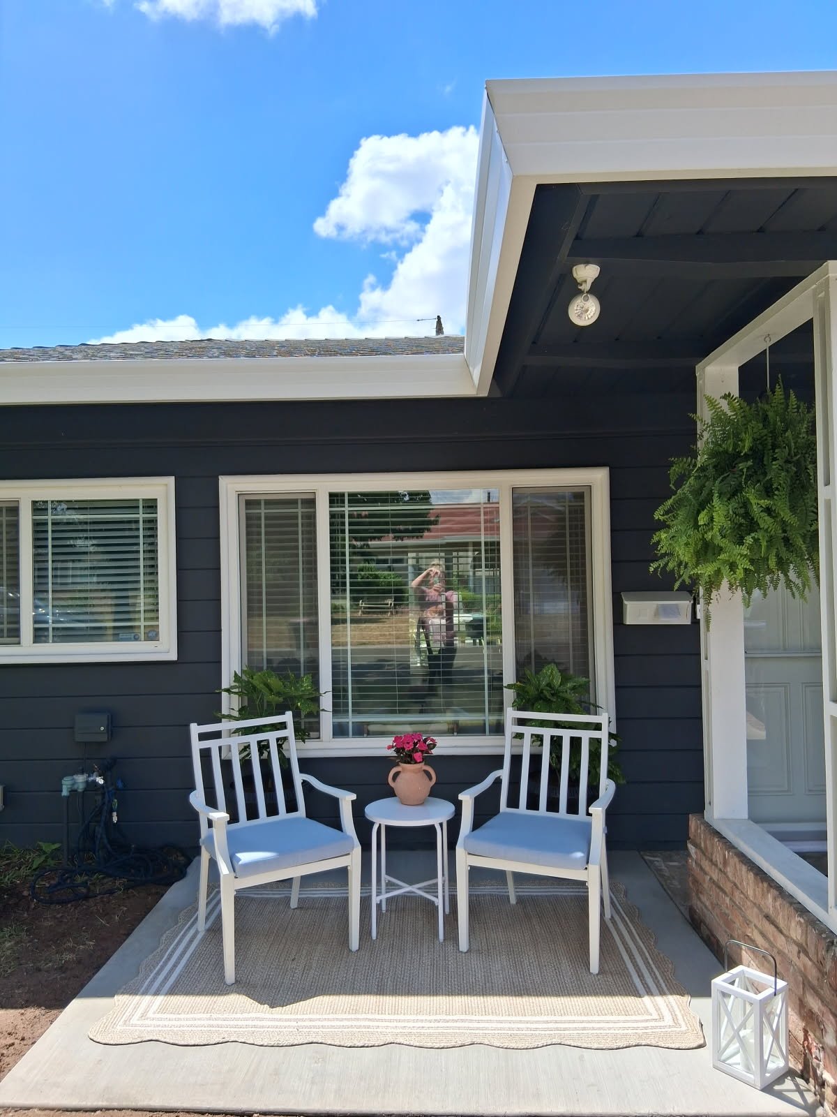 Patio area with two white chairs, a small white table, a vase with pink flowers, a beige rug, green plants, and a window with white blinds on a dark blue house exterior under a partly cloudy sky.