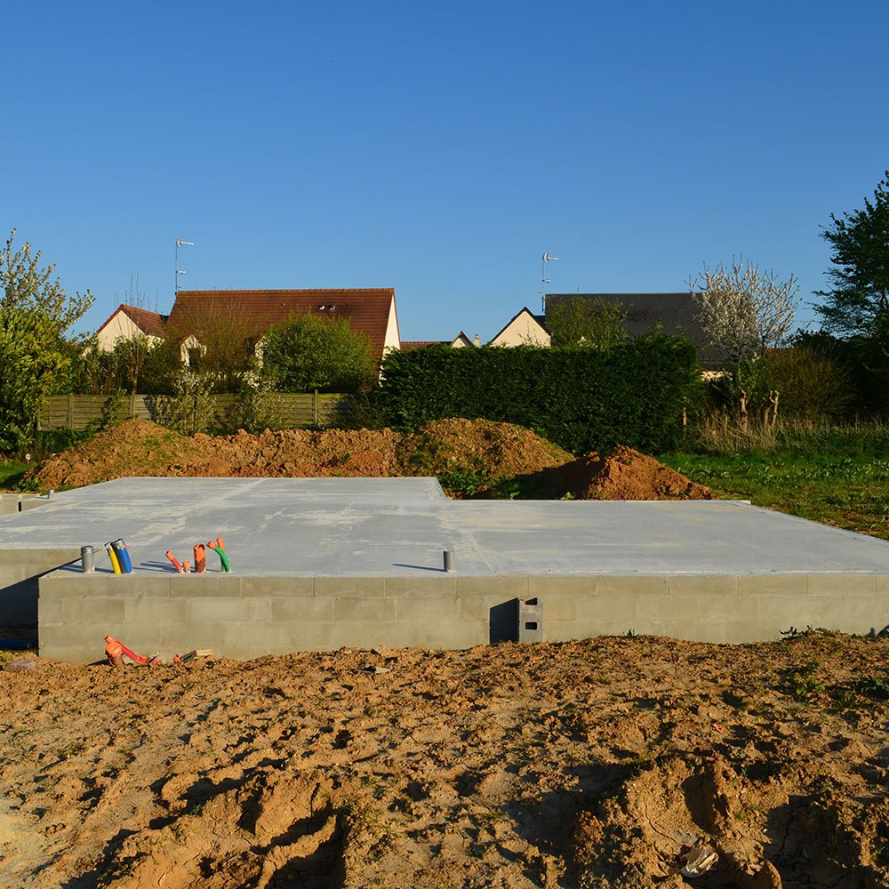 Construction site with concrete foundation in a residential area, dirt piles, and houses in the background under a clear blue sky.