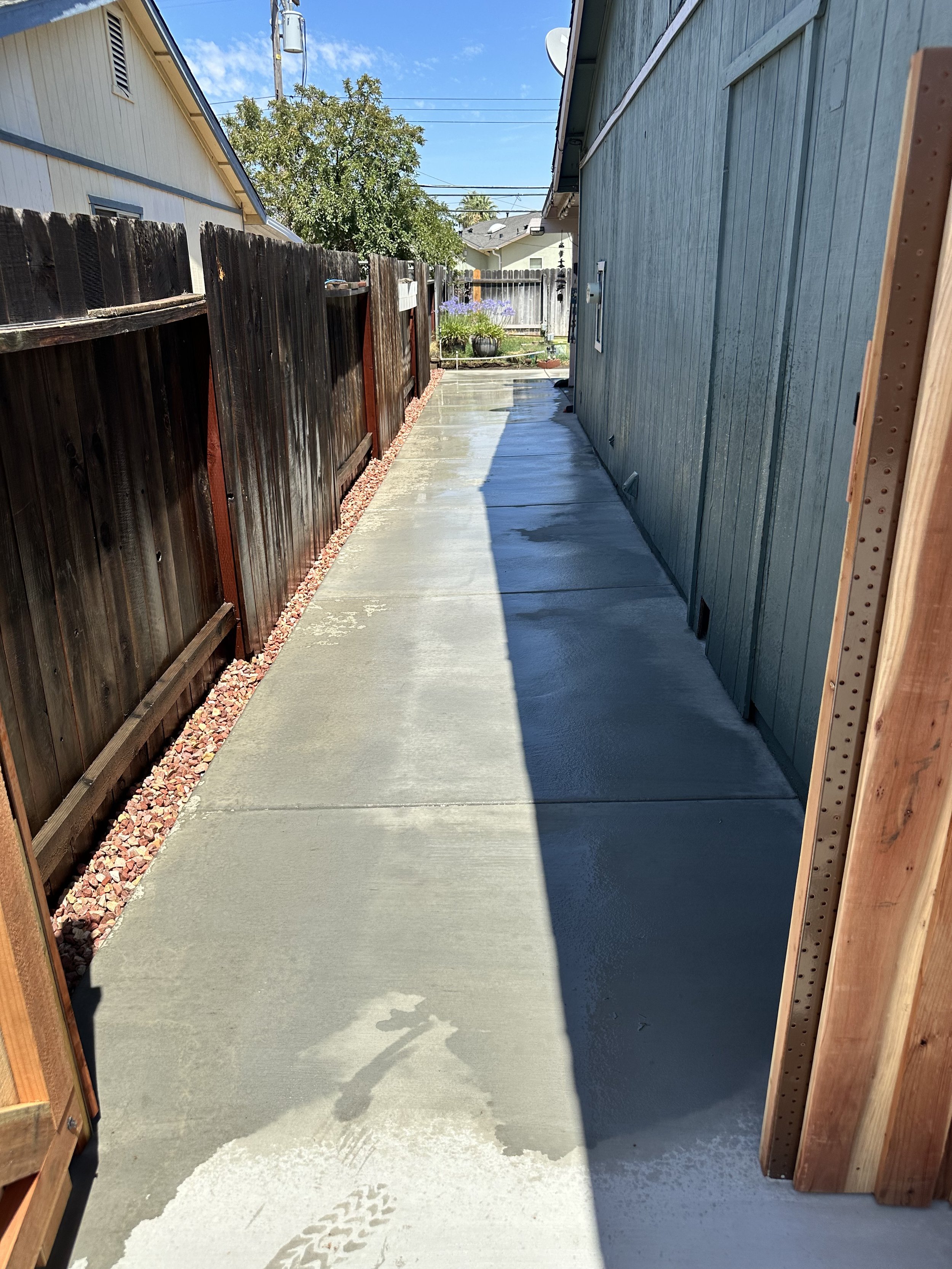 View of a narrow concrete sidewalk between a dark wooden fence and the side of a building, with some wet patches, on a sunny day.