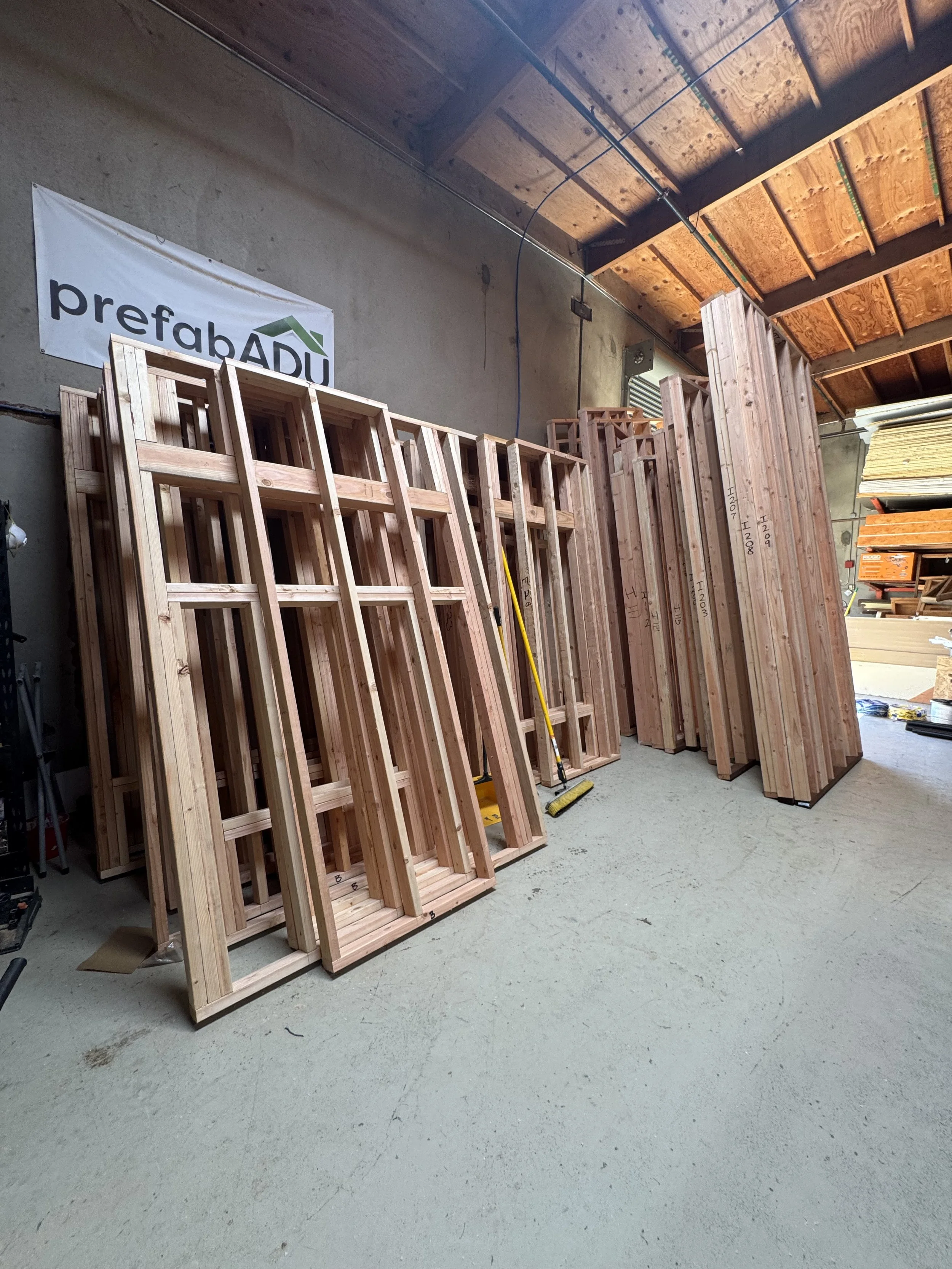 Stacks of wooden framing lumber in a warehouse with a 'prefabADU' sign on the wall.