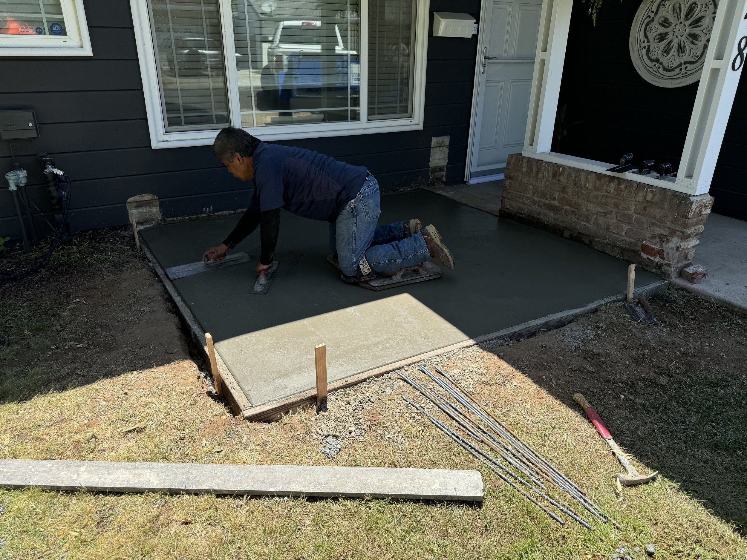 A person kneeling on a concrete slab, smoothing the surface with a trowel, in front of a house.