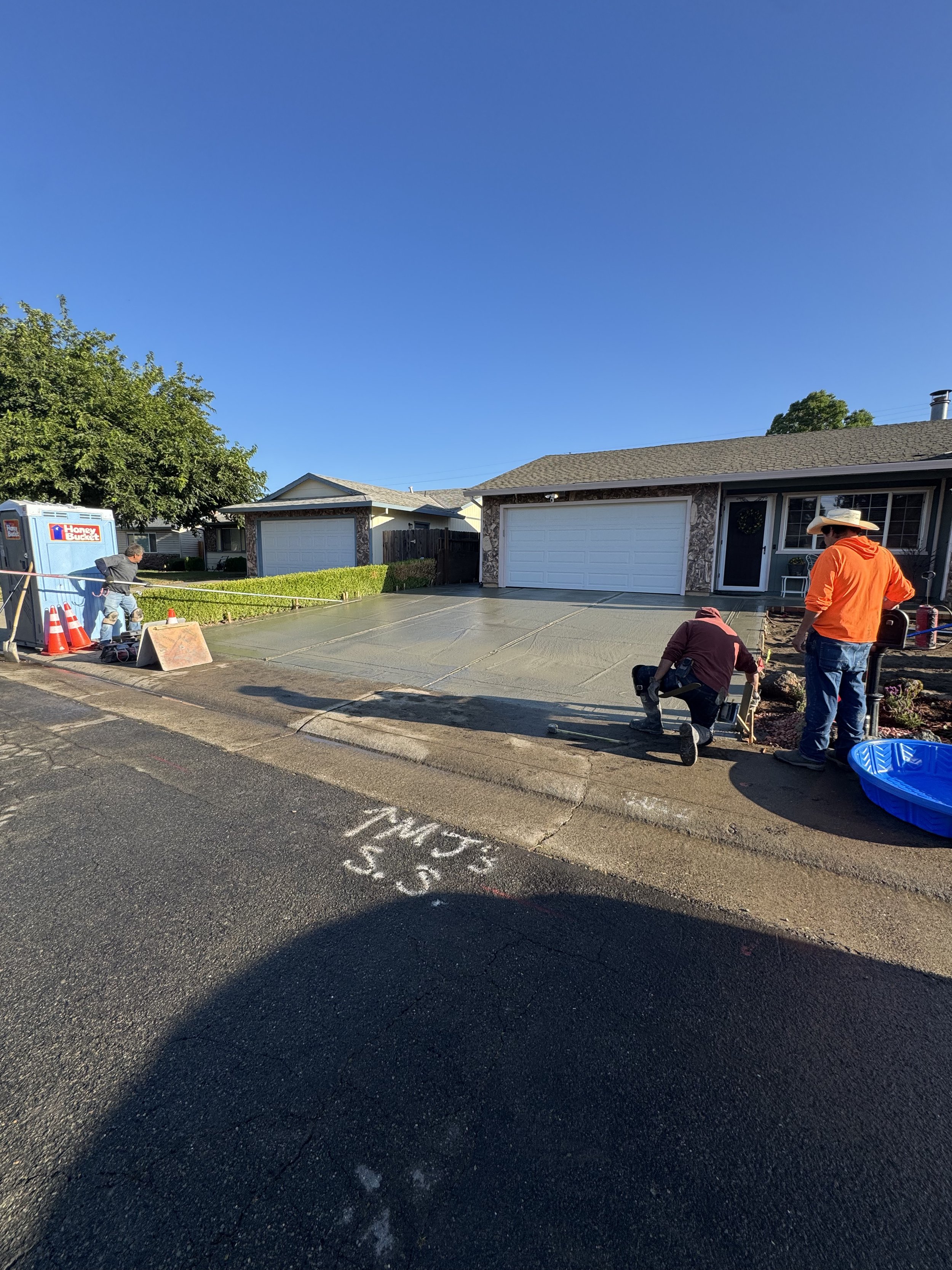 Three workers are pouring a fresh concrete driveway in front of a house on a sunny day, with a tree, neighboring houses, and a blue sky in the background.