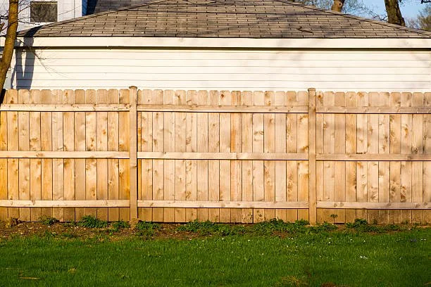 Wooden backyard fence in front of a house with a shingle roof and white siding, with green grass in the foreground.
