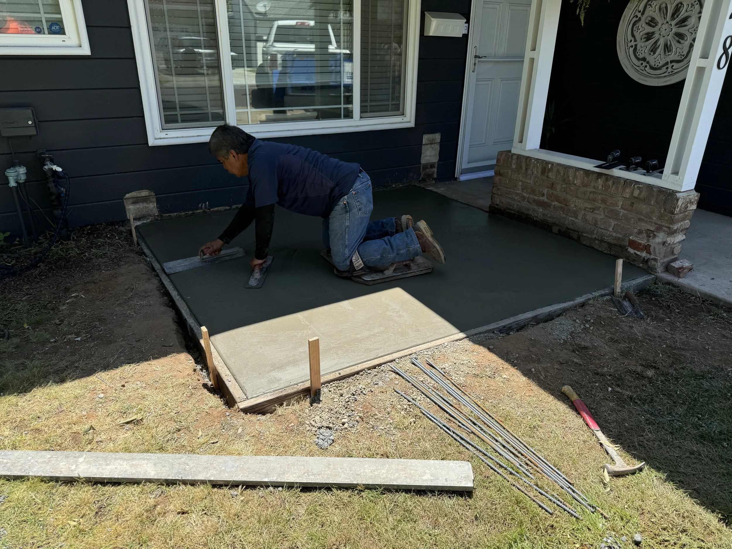 A person kneeling on a piece of cardboard, smoothing wet concrete with a trowel in front of a house, with tools and wooden stakes nearby.