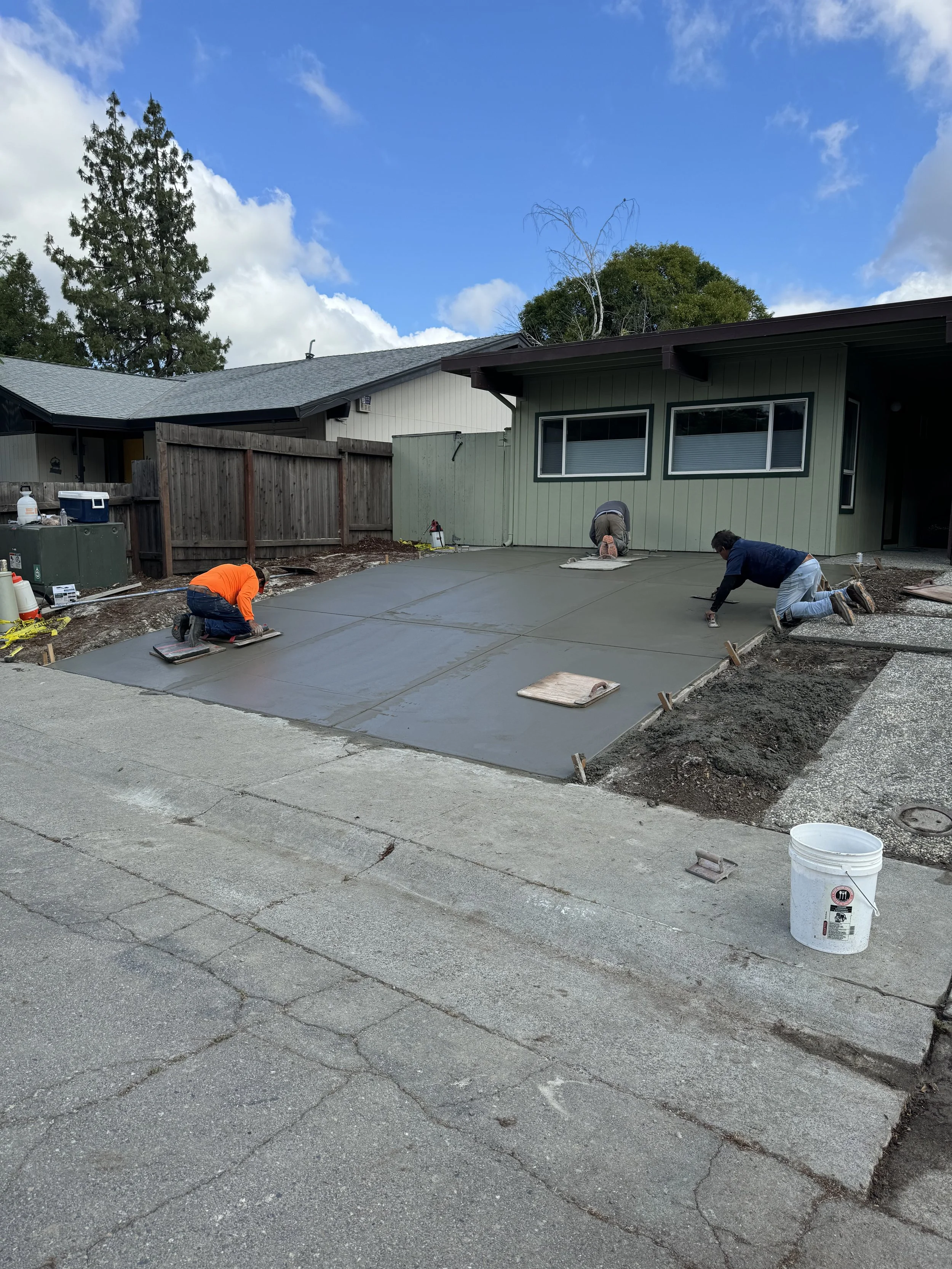 Three workers are pouring and smoothing concrete on a driveway in front of a green house, with a blue sky and clouds overhead.