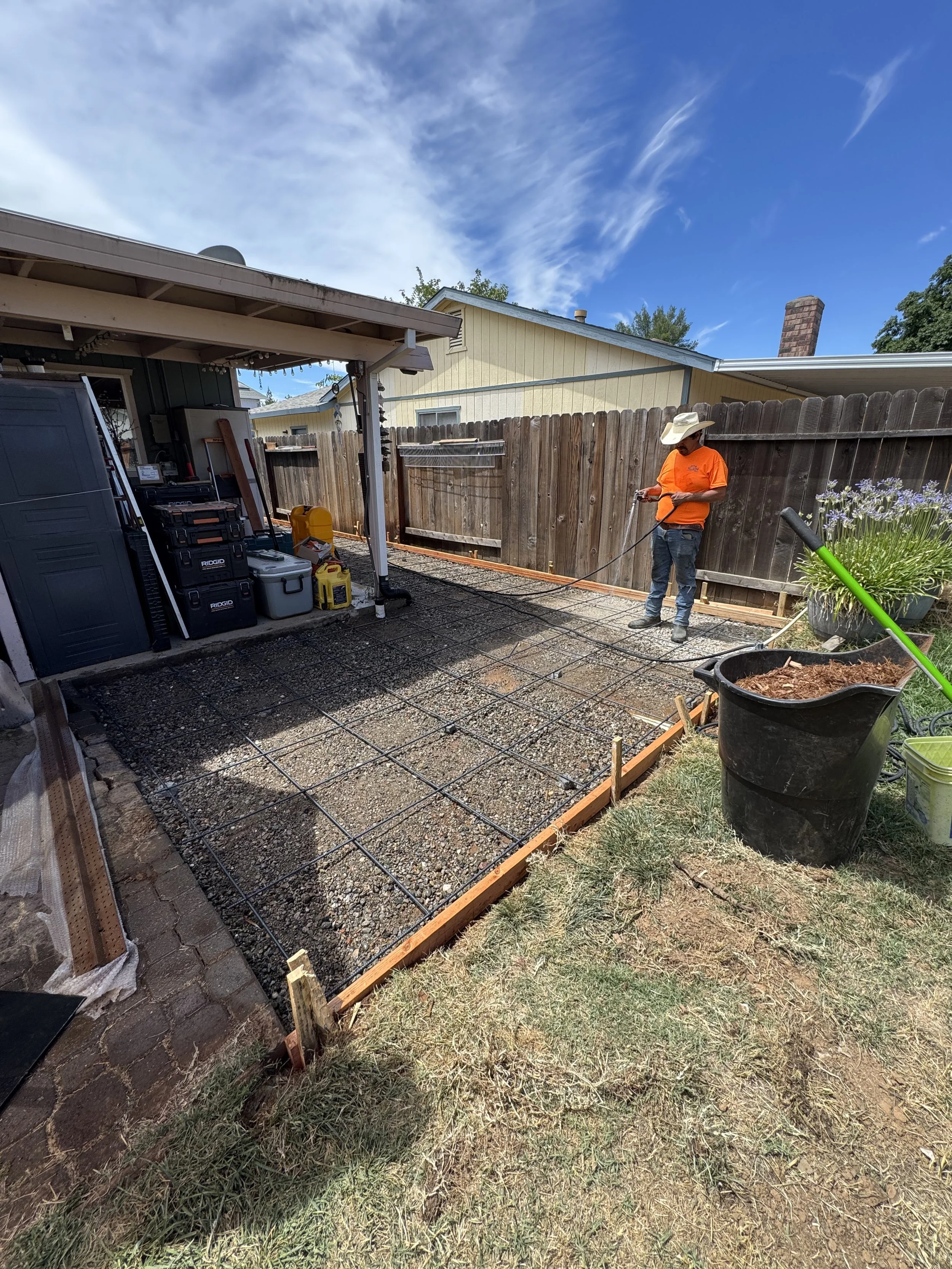 A person wearing an orange shirt and white hat is working on a backyard patio construction with steel reinforcement rebar. There are tools and containers nearby, and a wooden fence surrounds the area under a partly cloudy sky.