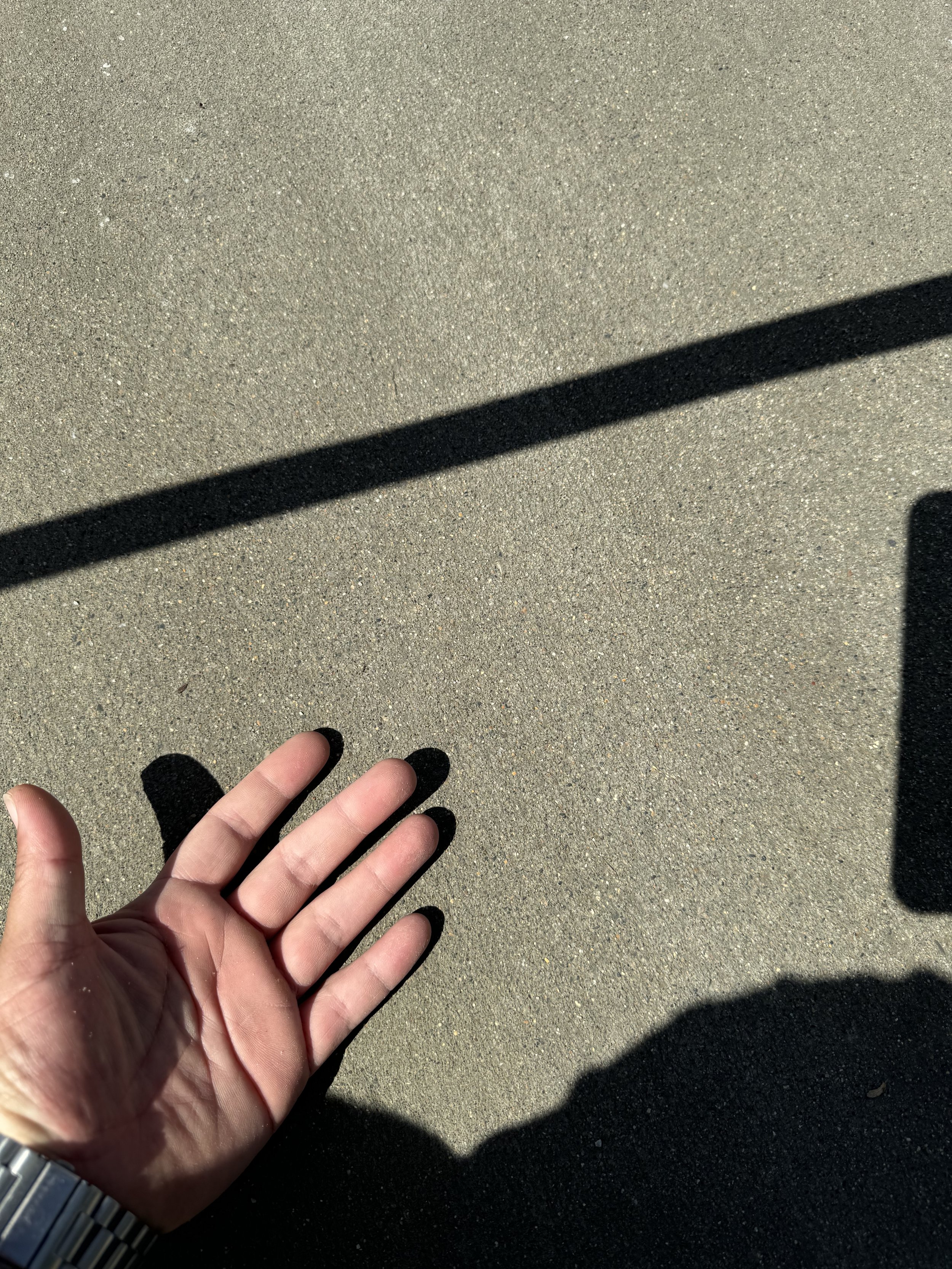 Photo of a person's left hand with a shadow, on textured pavement with shadows from a railing and a smartphone.