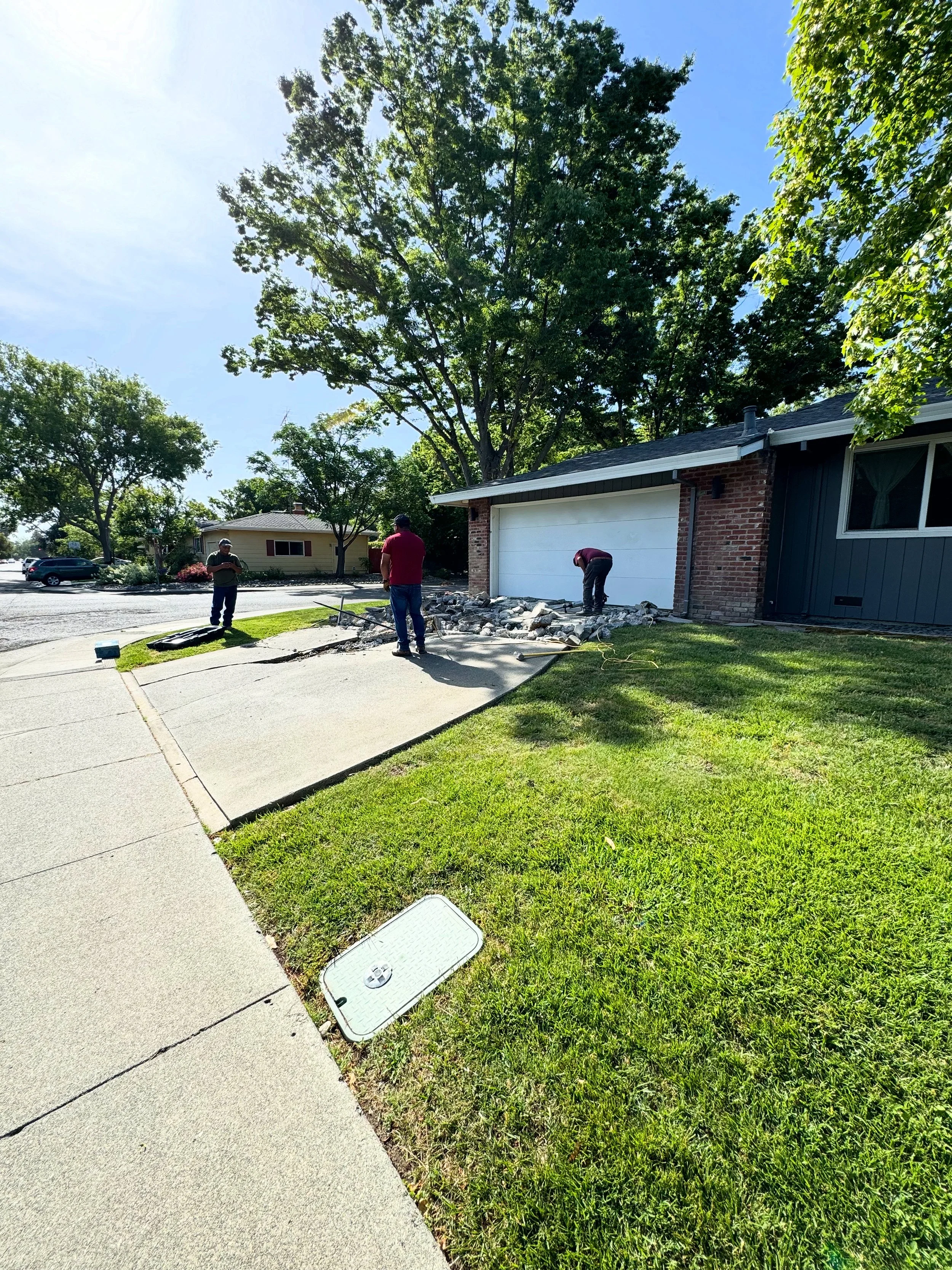 Three workers are replacing a section of concrete driveway in front of a house, with rocks and tools around.