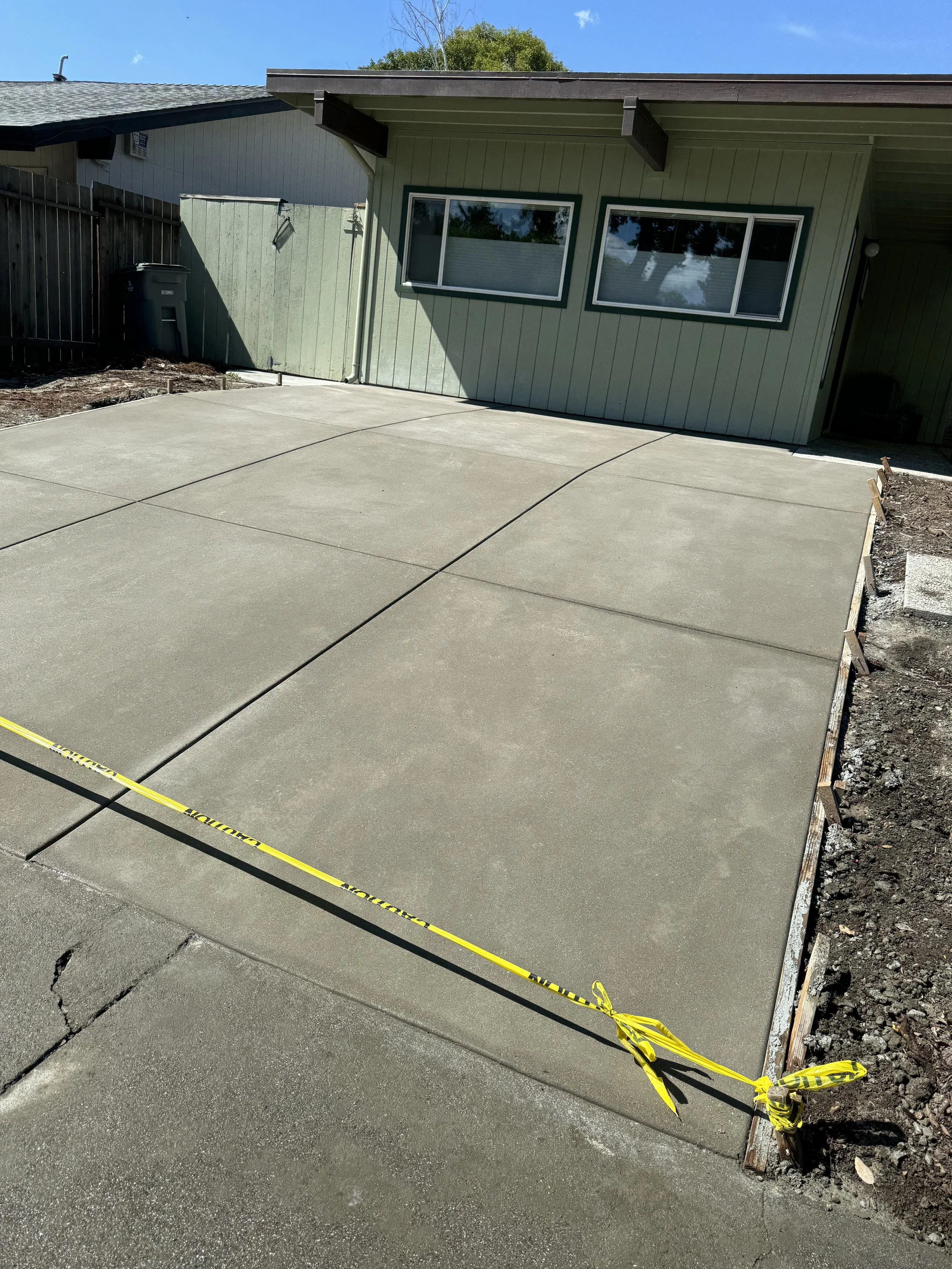 Freshly poured concrete driveway in front of a house with two windows, a green exterior, and a brown roof. A yellow caution tape is stretched across part of the driveway, and there is some soil and yard work around the edges.