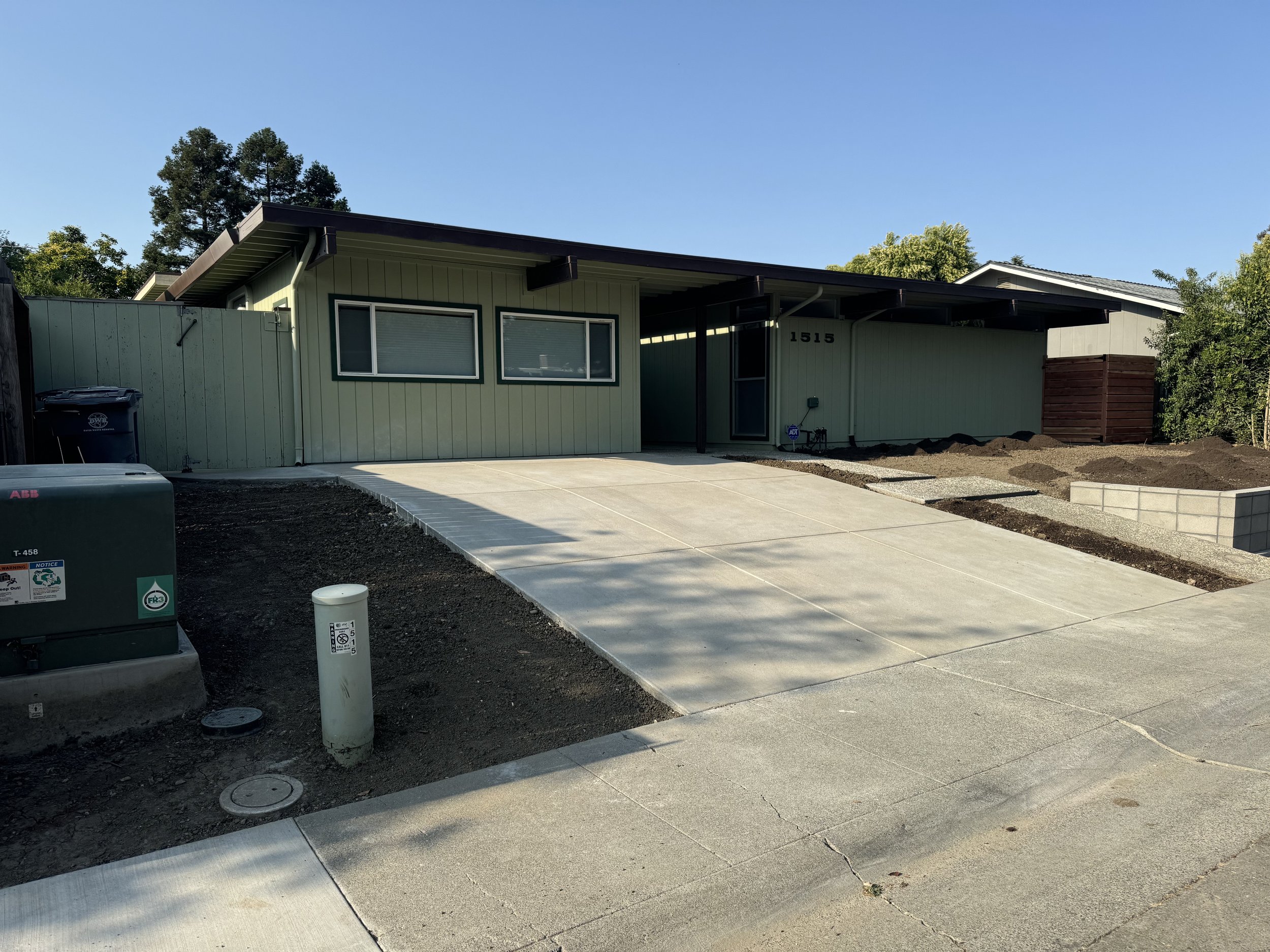 A modern single-story house with a green exterior, two large front windows, a concrete driveway, and a front yard under landscaping. The house has house number 1515 displayed near the door, and the sky is clear with a few trees in the background.