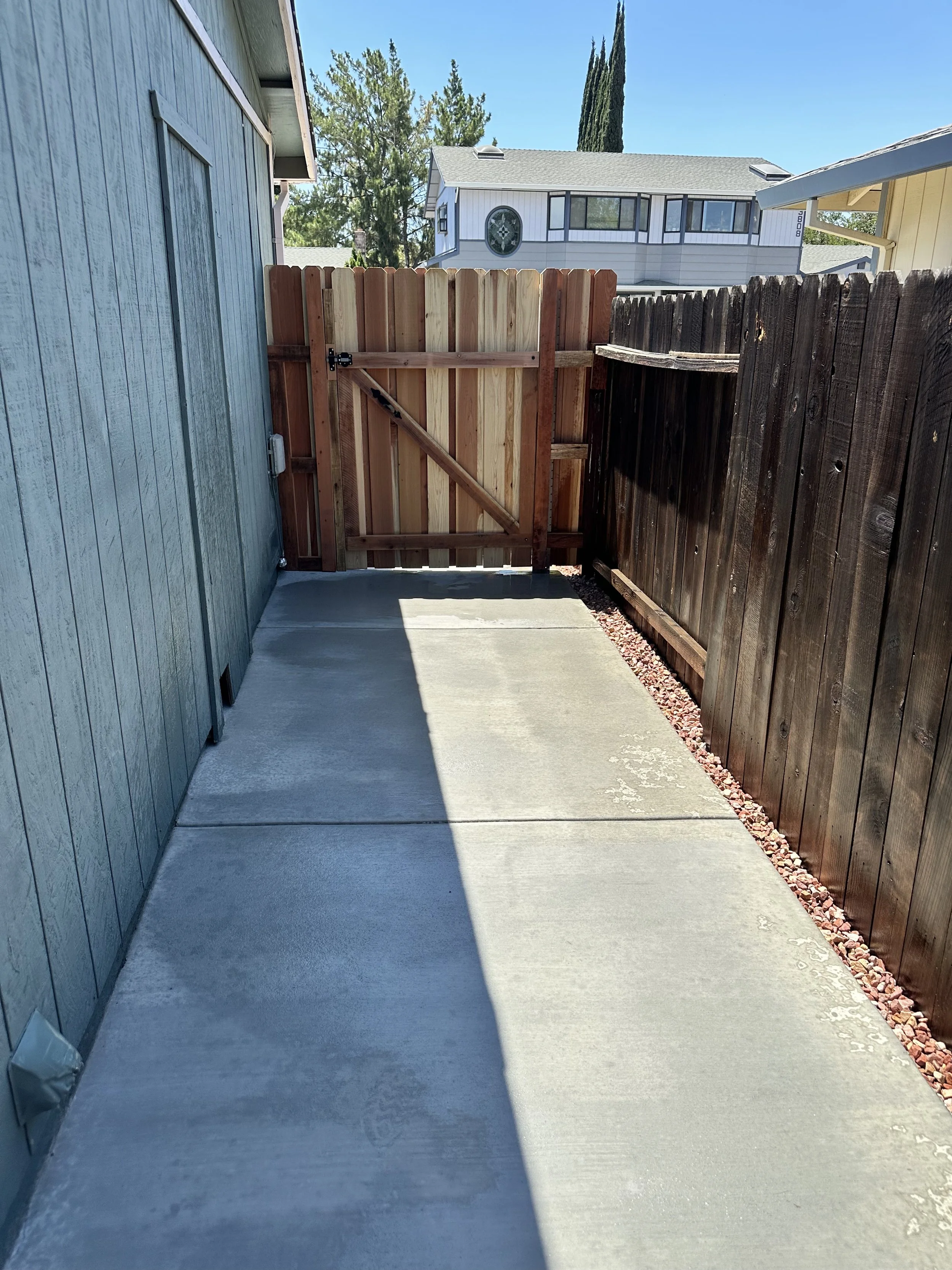 Empty concrete backyard patio with wooden fence gate at the end, shadow cast on the ground, neighboring houses visible in the background.