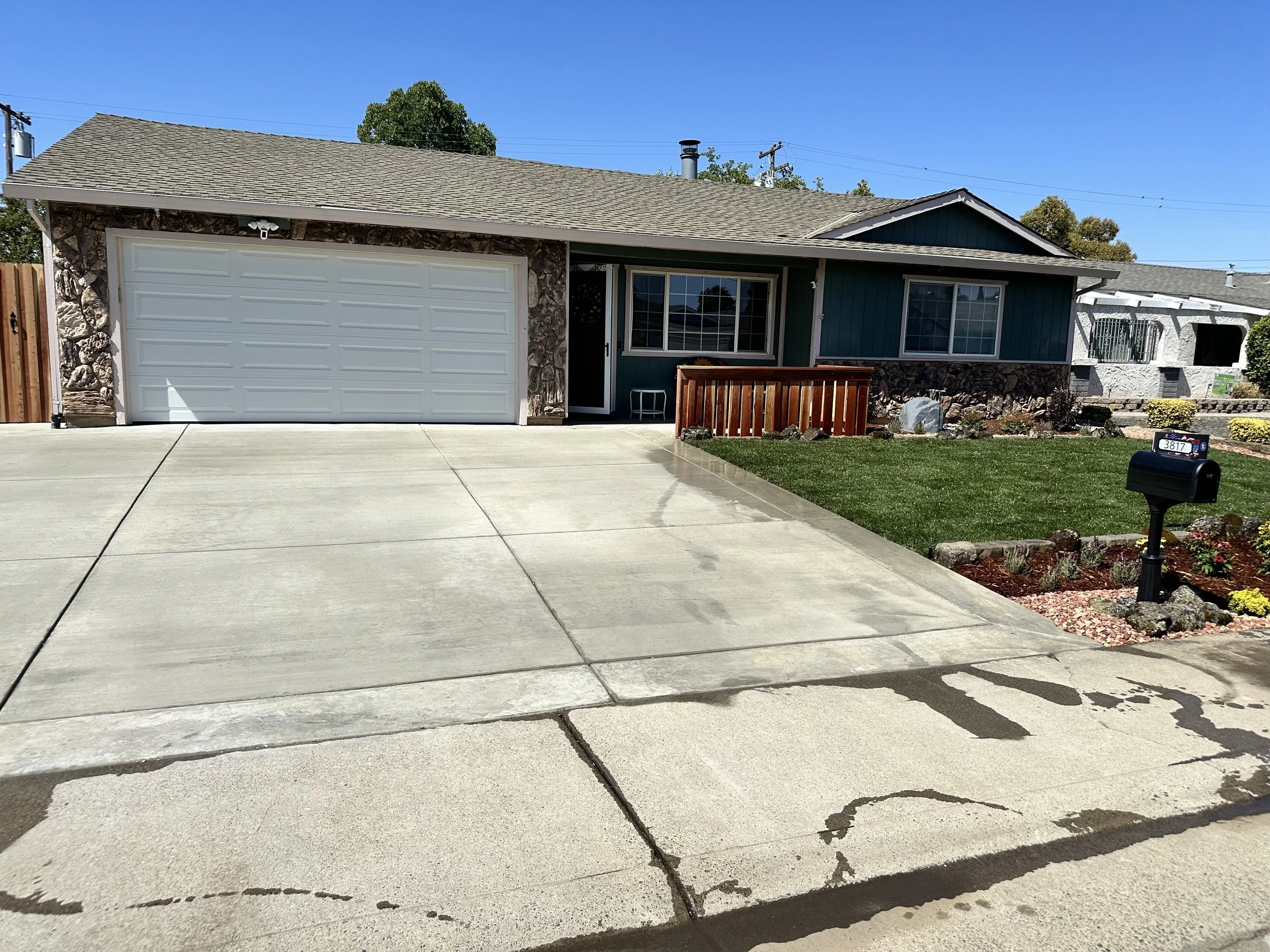 Front view of a house with a concrete driveway, green grass lawn, mailbox, and a wooden fence around the porch. The house has a two-car garage, teal siding, stone accents, and large windows.