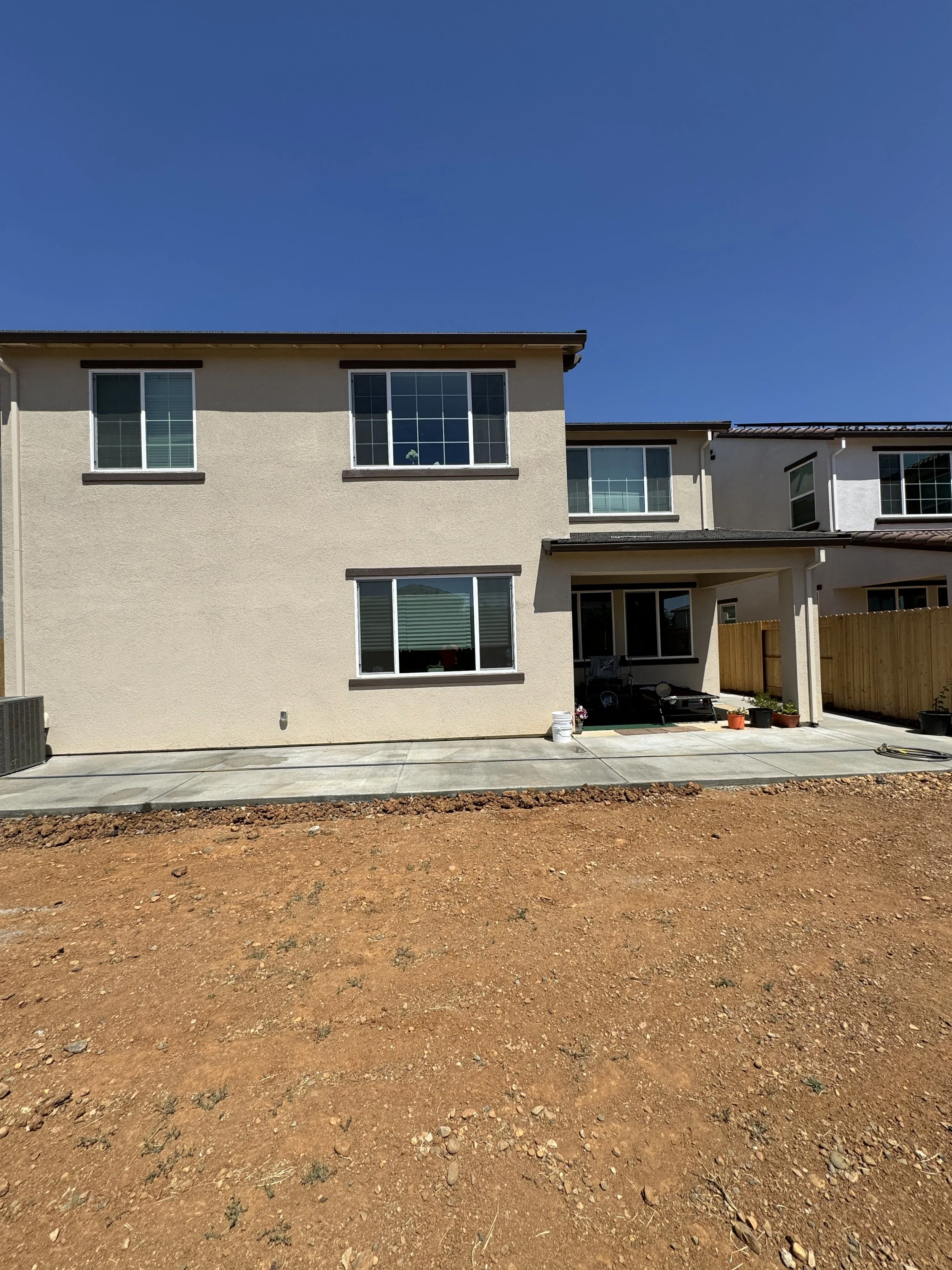 Back exterior of modern two-story house with a small patio, gardening supplies, and a fenced yard at the edge of a landscaped lawn under clear blue sky.