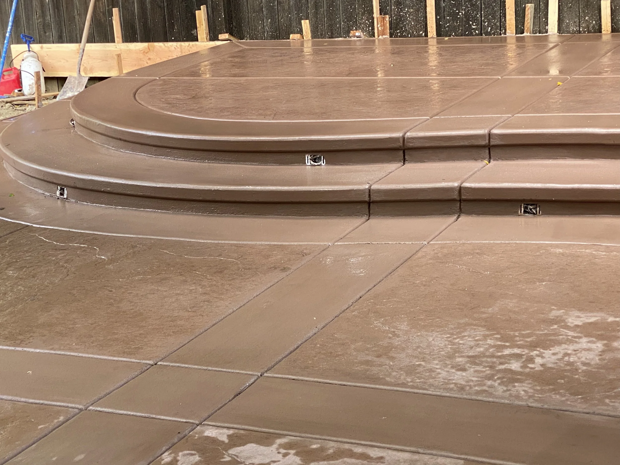 Brown concrete steps leading to a deck under construction in a backyard with a fire extinguisher and garden tools visible in the background.
