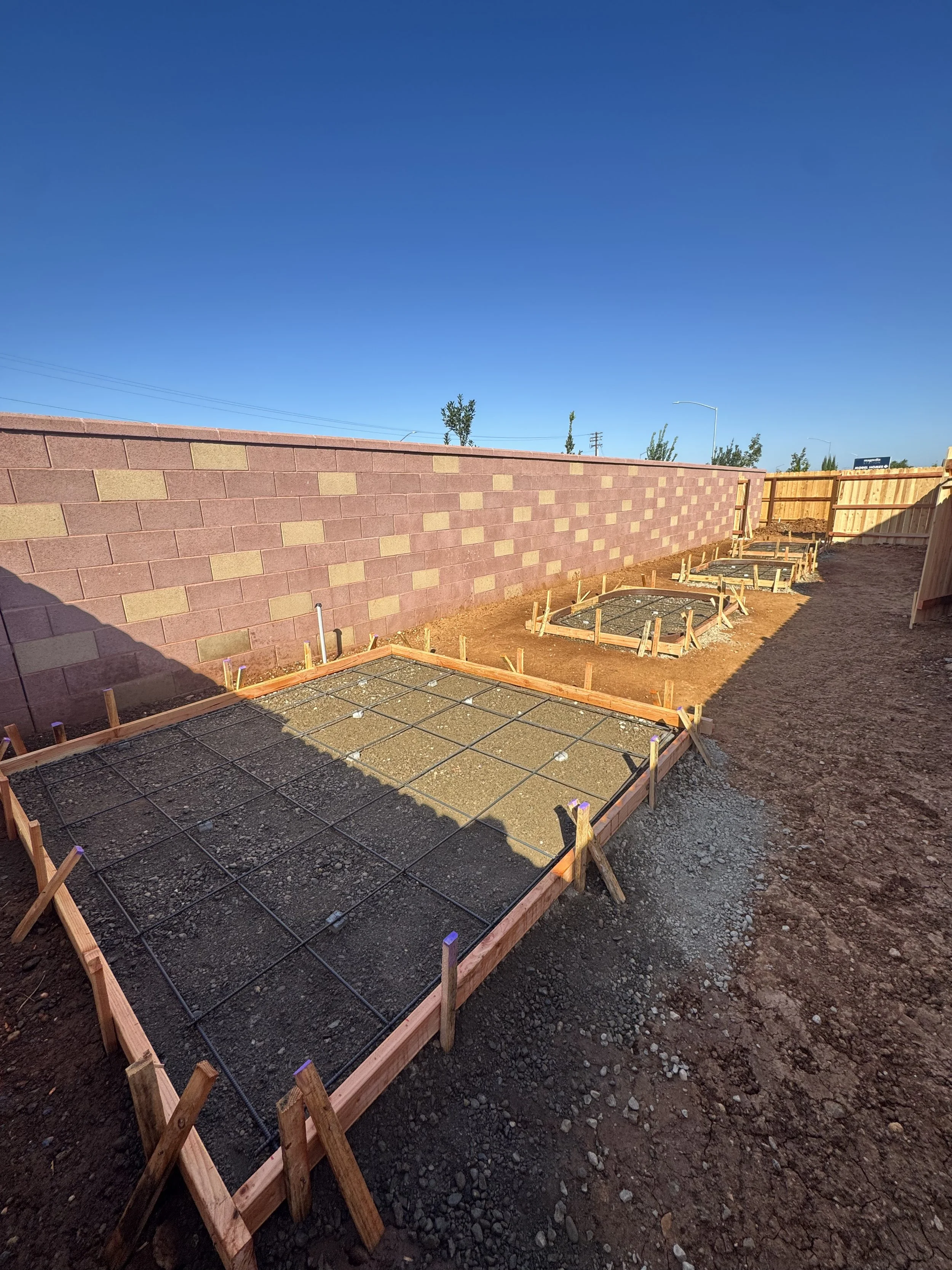 Construction site with wooden frames and rebar for concrete pouring, along a brick wall and a clear blue sky.