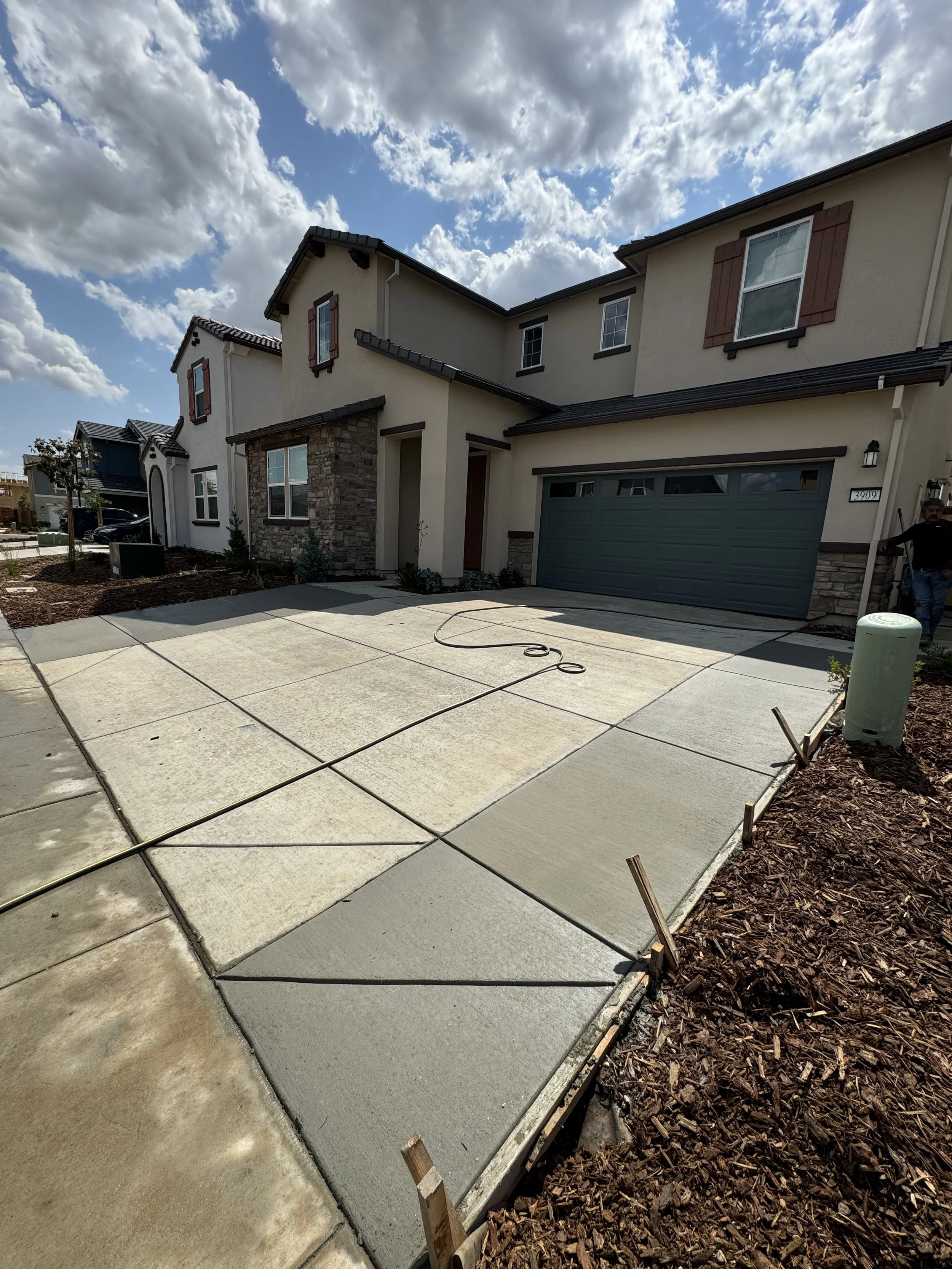 Newly poured concrete sidewalk in front of a modern two-story house with a gray garage door, stone accents, and brown window shutters. Construction tools and materials are visible.
