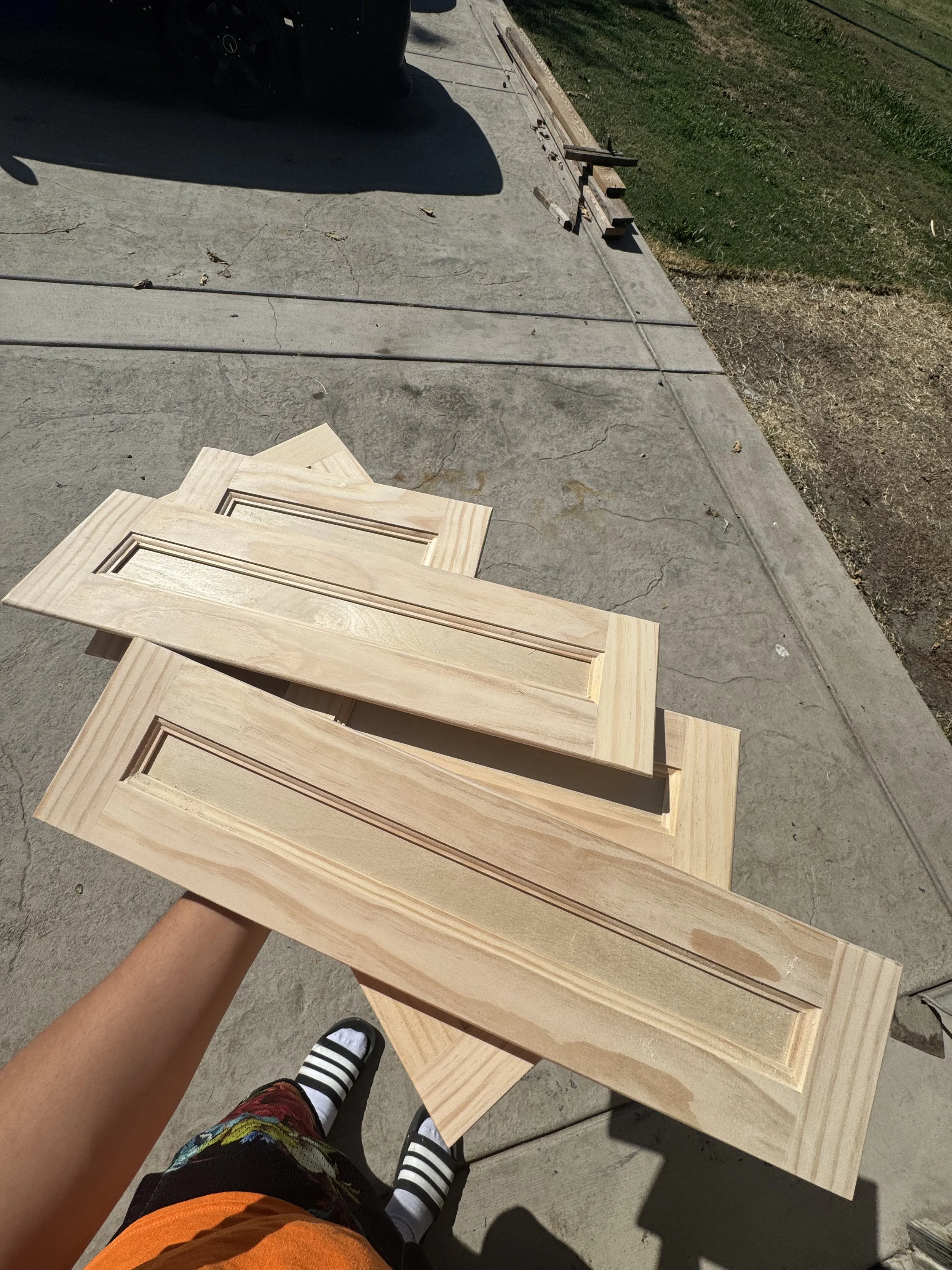 Person holding three unfinished wooden cabinet doors outdoors on a concrete surface.