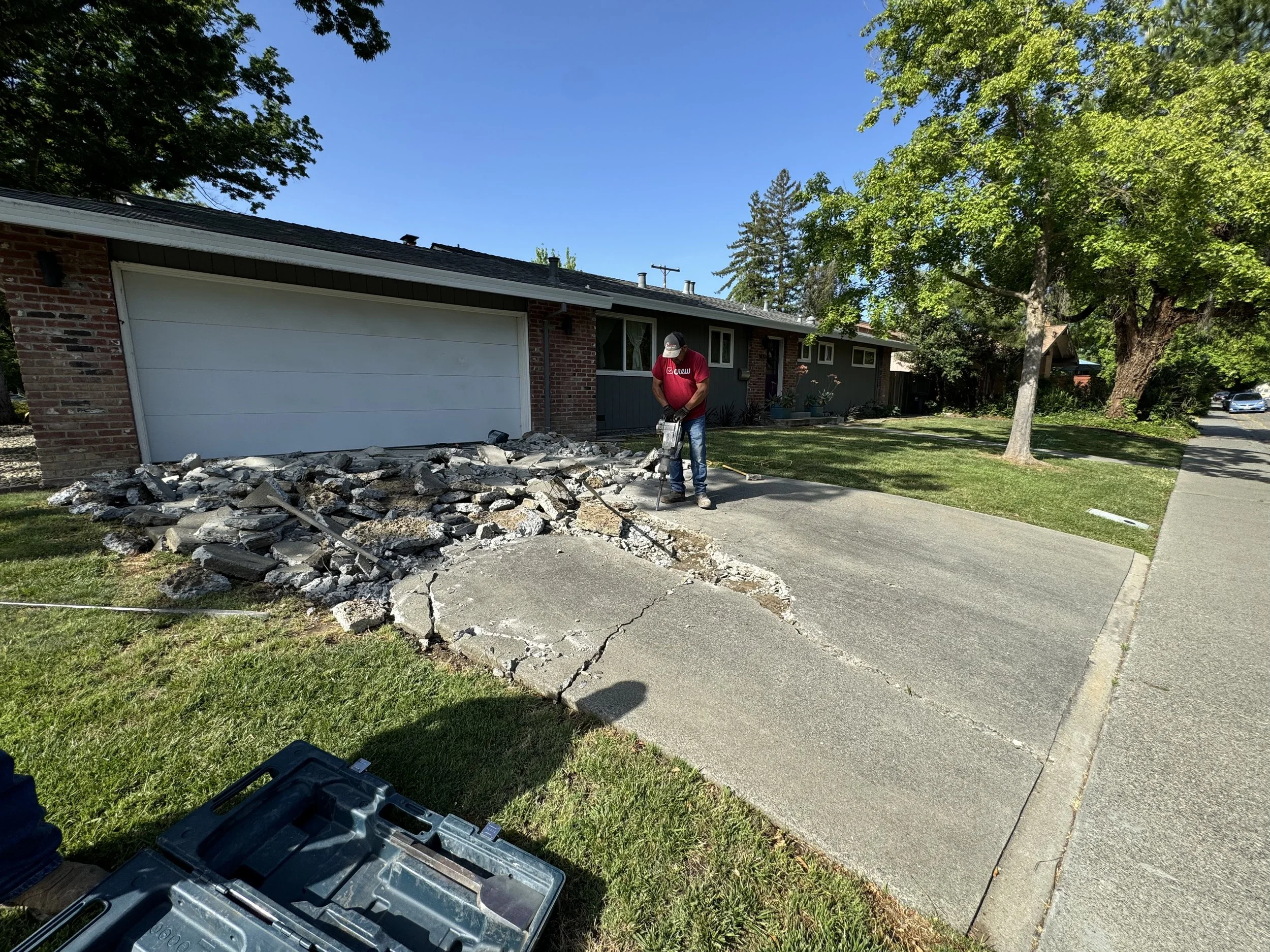 A person in a red shirt and cap using a jackhammer to break up a concrete driveway in front of a house, with a pile of broken concrete and debris nearby.