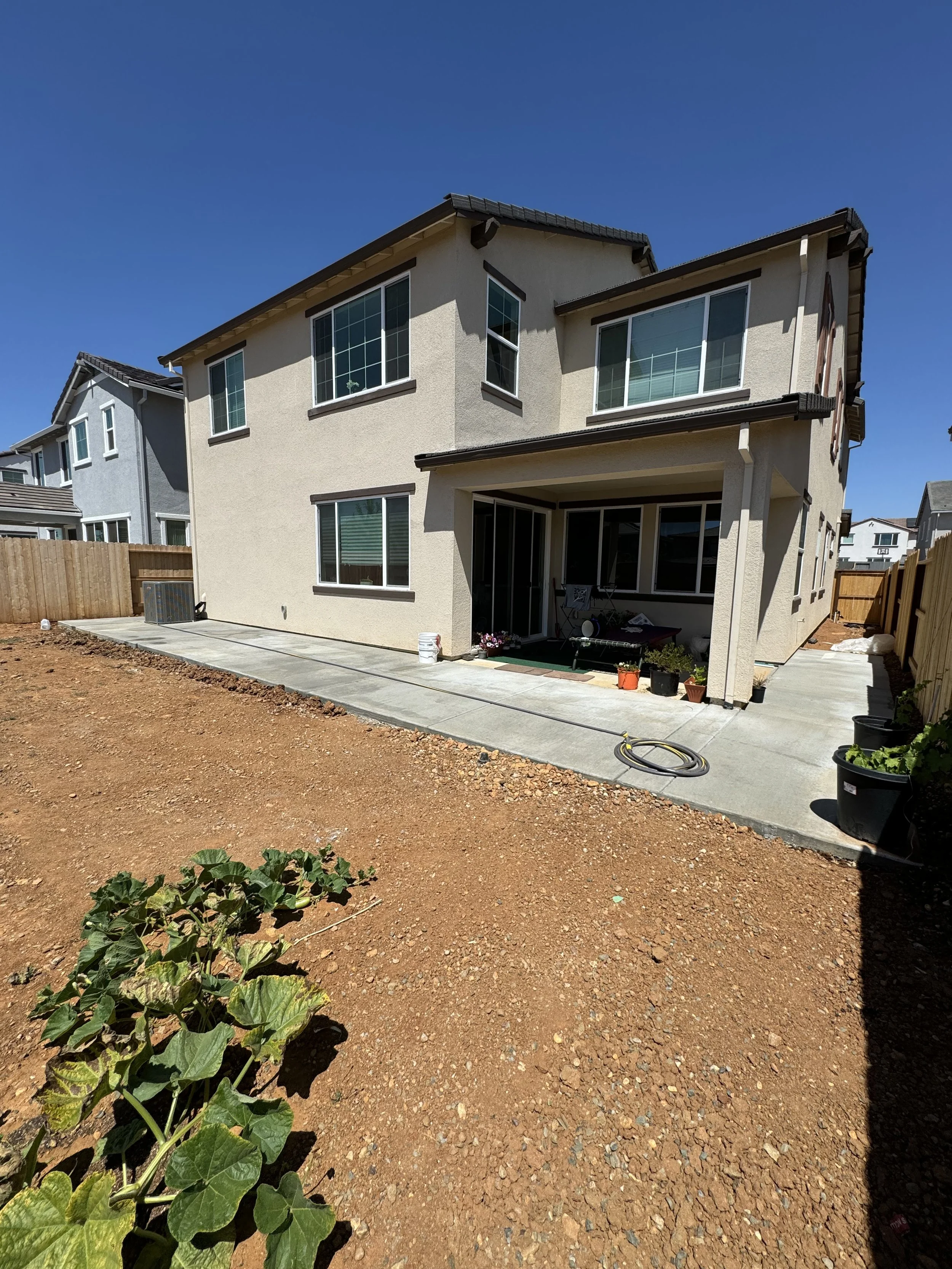Backyard of a two-story house with a concrete patio and a dirt yard. The house has beige stucco siding, several windows, and a covered patio area with potted plants and outdoor furniture. Wooden fencing surrounds the yard, and there are neighboring h