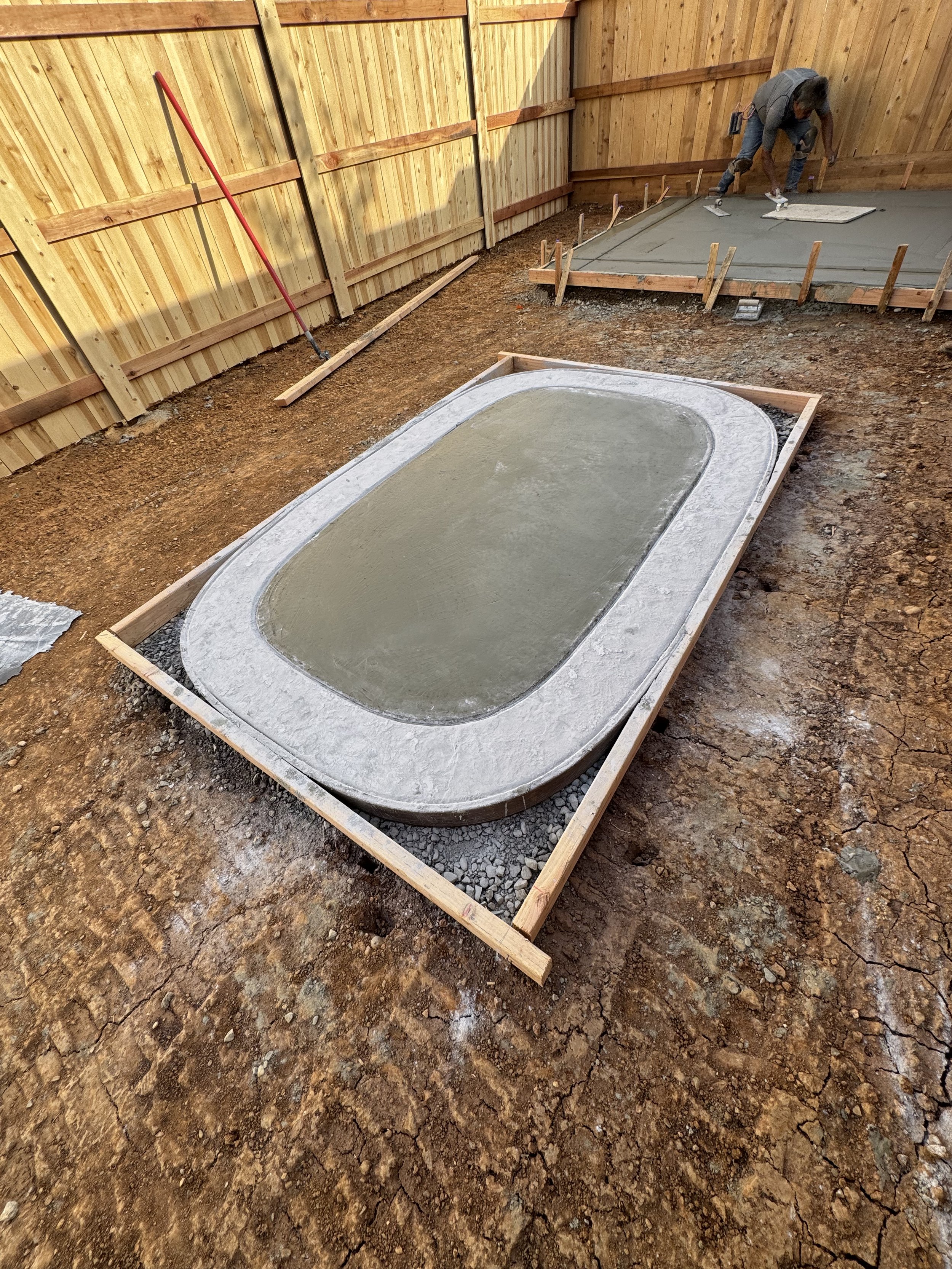 Construction site with a concrete pool in the process of being built, surrounded by a wooden frame, with workers in the background pouring concrete.