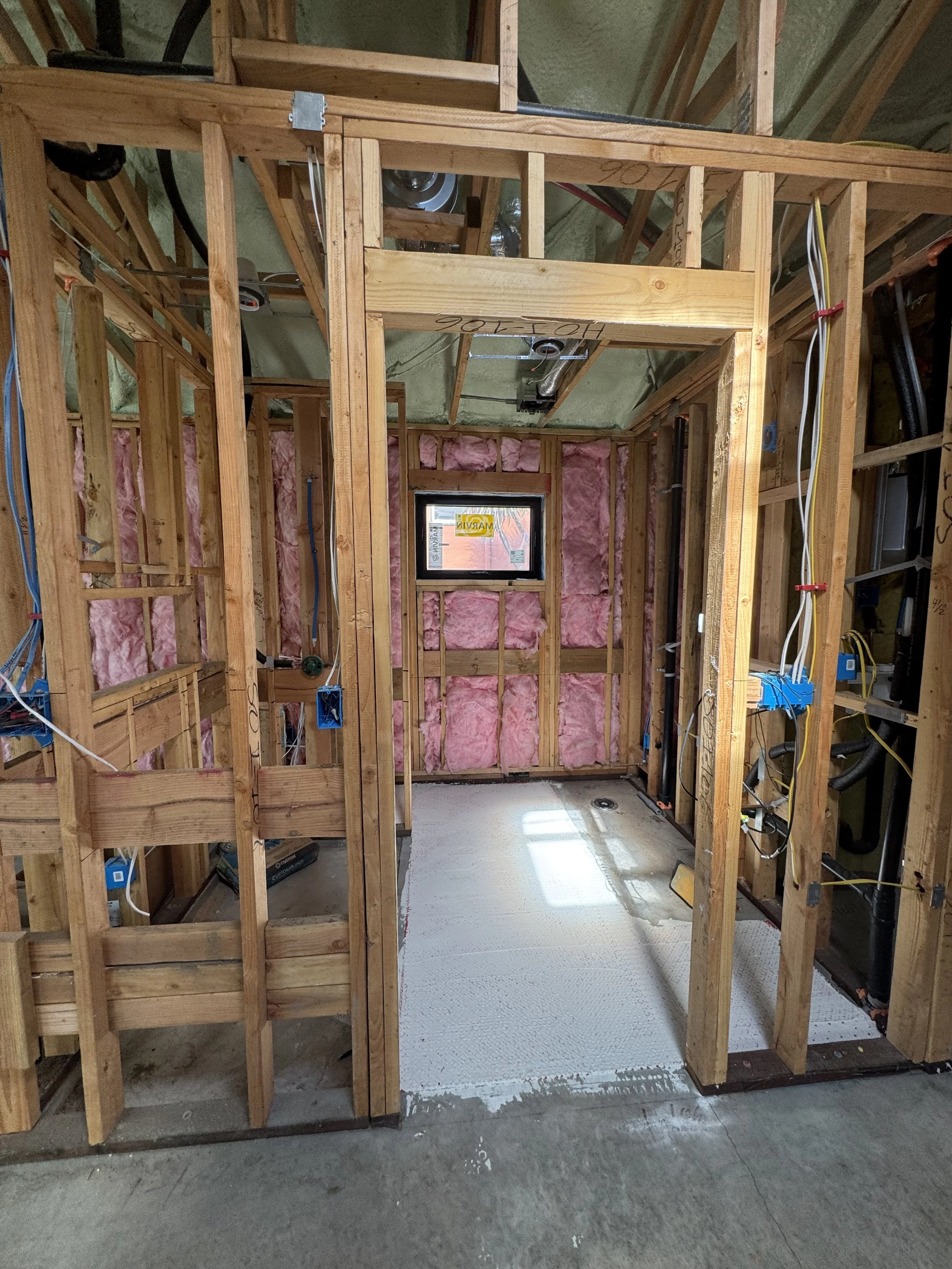 This image shows the framing of a small room under construction, with exposed wooden studs, pink insulation on the walls, and some electrical wiring. There is a small window on the back wall and a gray floor surface.